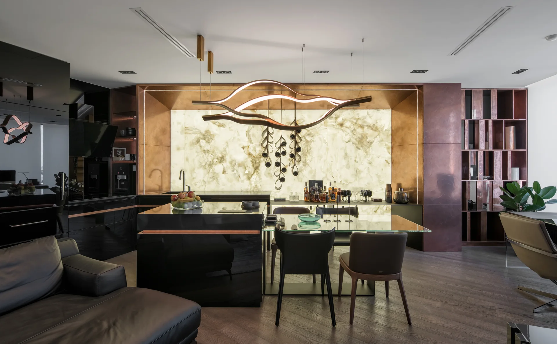 A modern kitchen and dining area featuring a black glossy island with an attached glass table set against a glowing backlit stone backsplash.