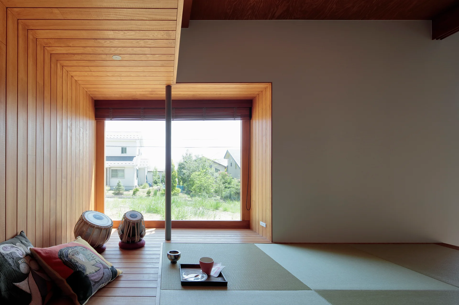 A minimalist room featuring traditional tatami floor mats, a large window framed in warm wood paneling, and decorative floor cushions.