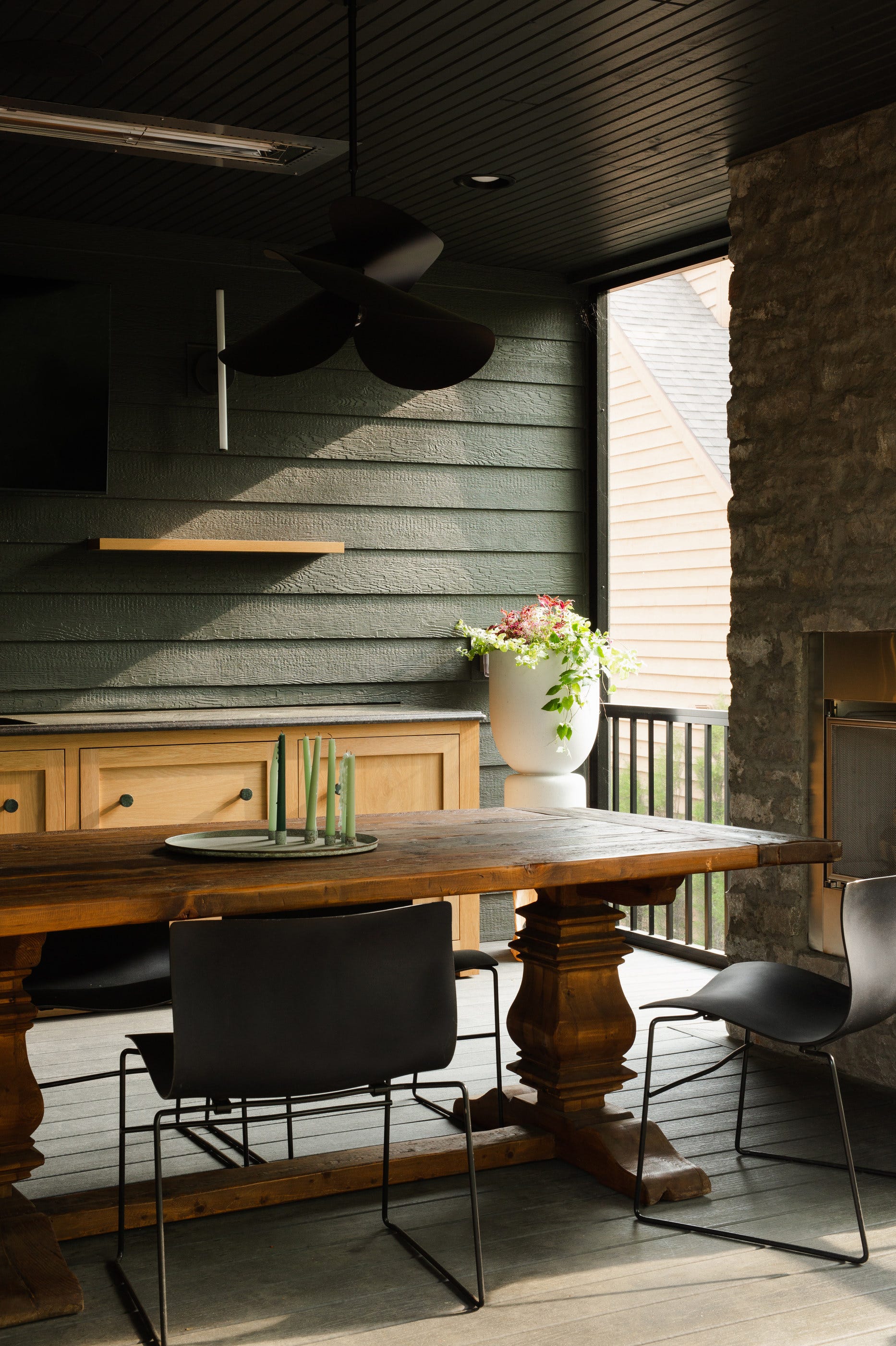 A screened-in porch features a rustic wooden dining table, black chairs, a stone fireplace, and dark horizontal wood siding.