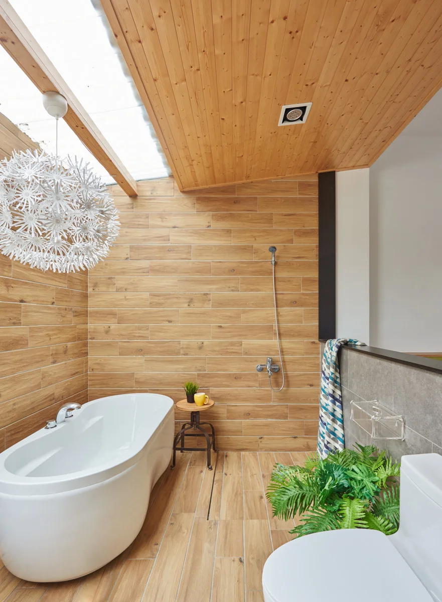 A modern bathroom featuring wood-look plank wall and floor tiles, a white freestanding bathtub under a skylight, and a large white spiky pendant light.