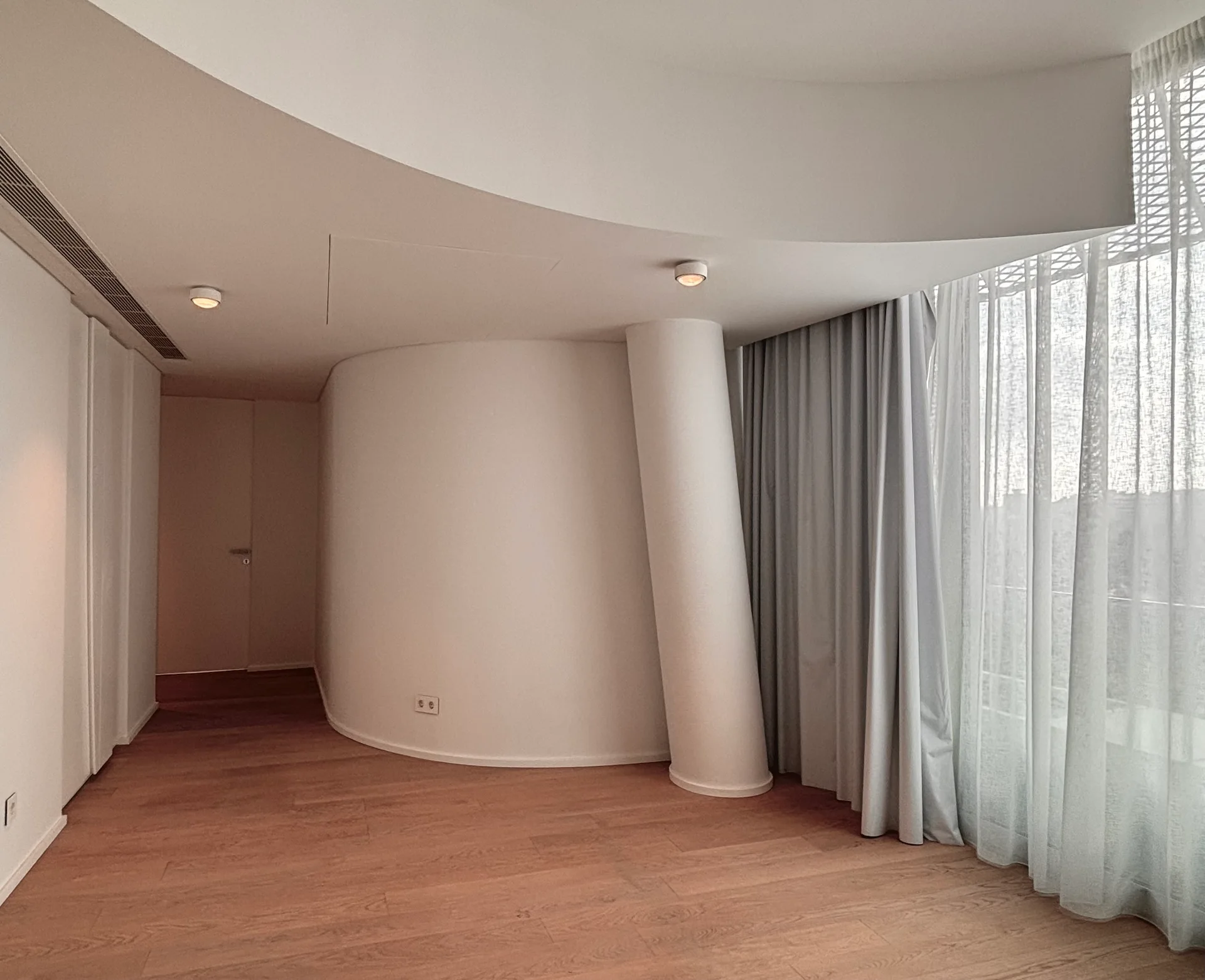An empty minimalist room featuring a smooth curved white partition wall, an angled column, light wood flooring, and floor-to-ceiling sheer grey curtains.