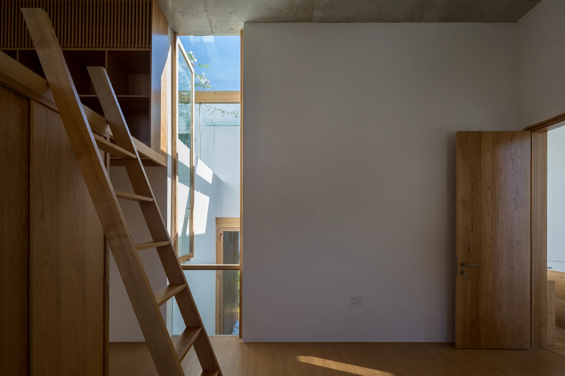A minimalist bedroom features a wooden loft ladder, built-in cabinetry, a plain white wall, and a vertical light well revealing the sky.