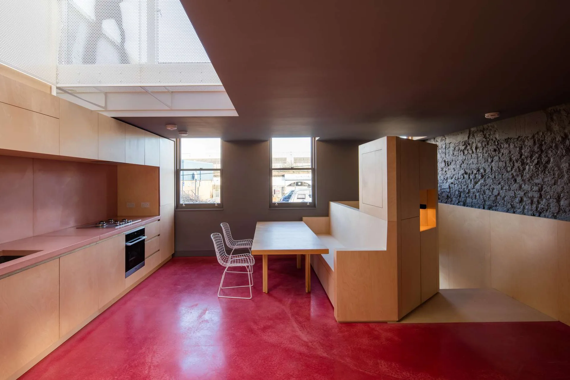 A minimalist kitchen with a bright red floor, light plywood cabinetry, a built-in dining nook, and a textured stone wall.