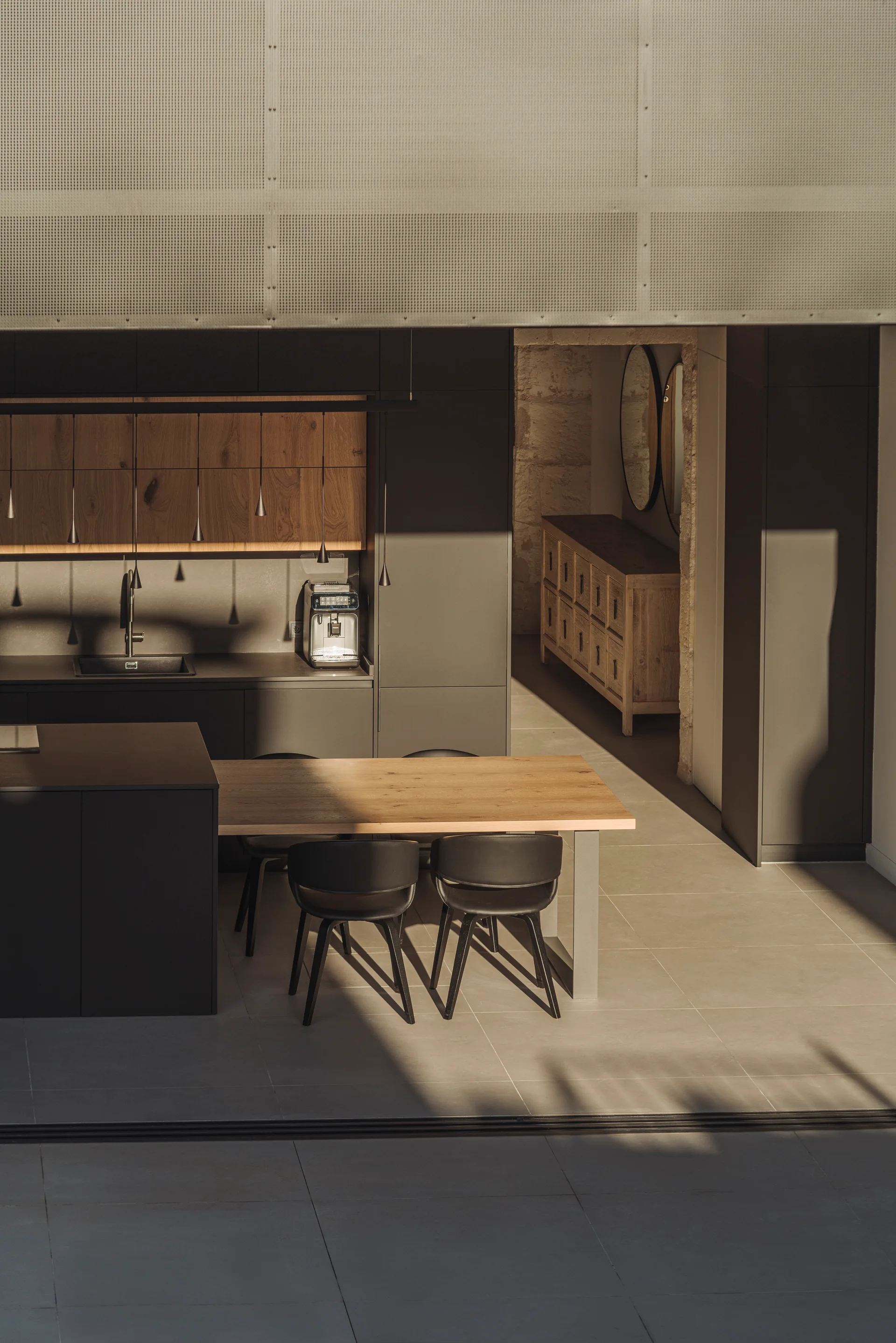 A minimalist charcoal kitchen features a matte dark island integrated with a light wood dining table, black chairs, and thin hanging pendant lights.