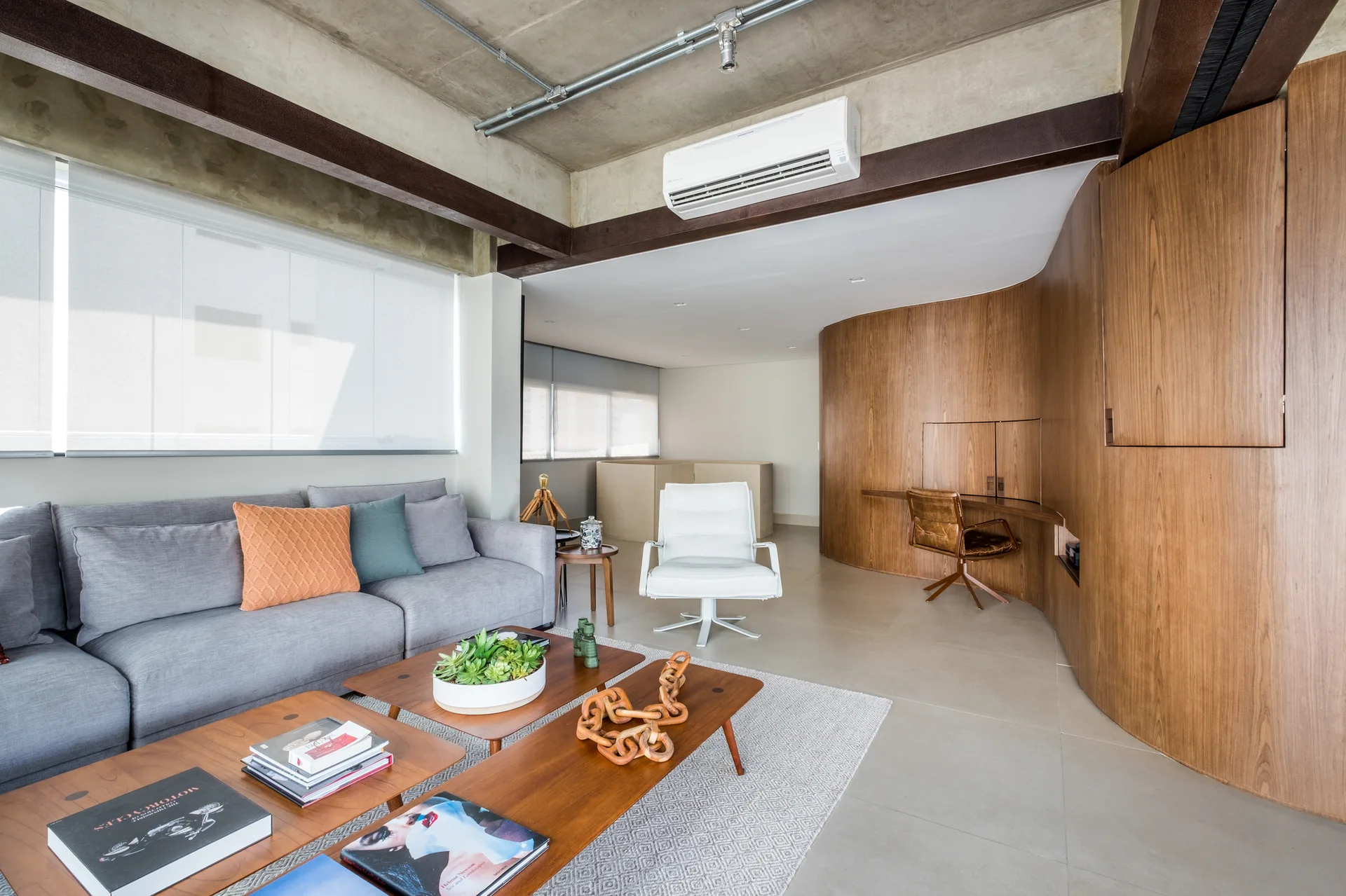A grey sofa sits across from curved wood wall paneling with an integrated desk under an exposed concrete ceiling in a modern living space.