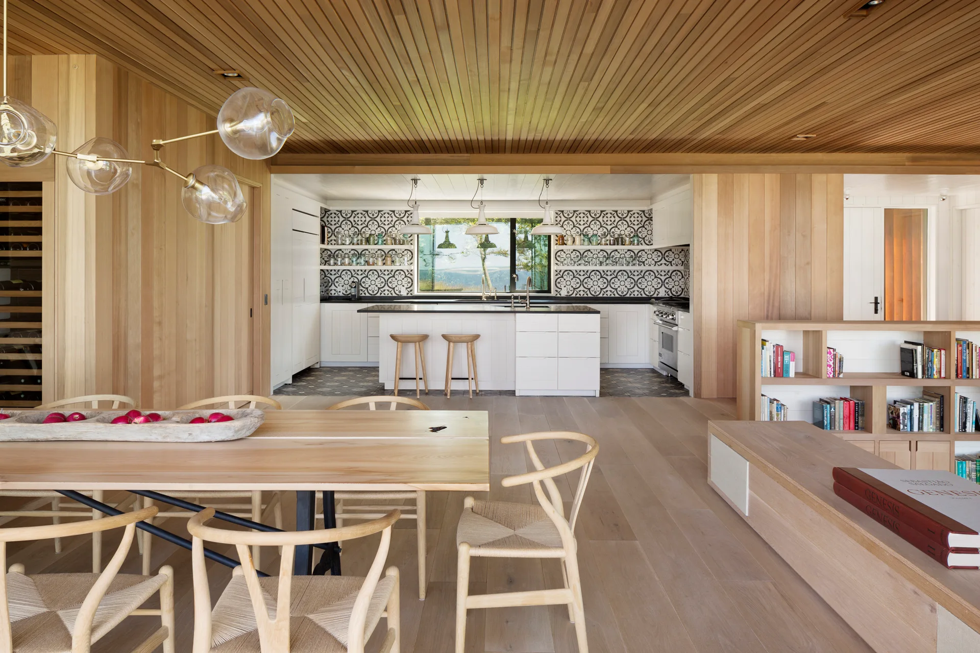 An open-concept dining space and kitchen featuring light wood paneling, wishbone chairs around a dining table, and a patterned tile backsplash.