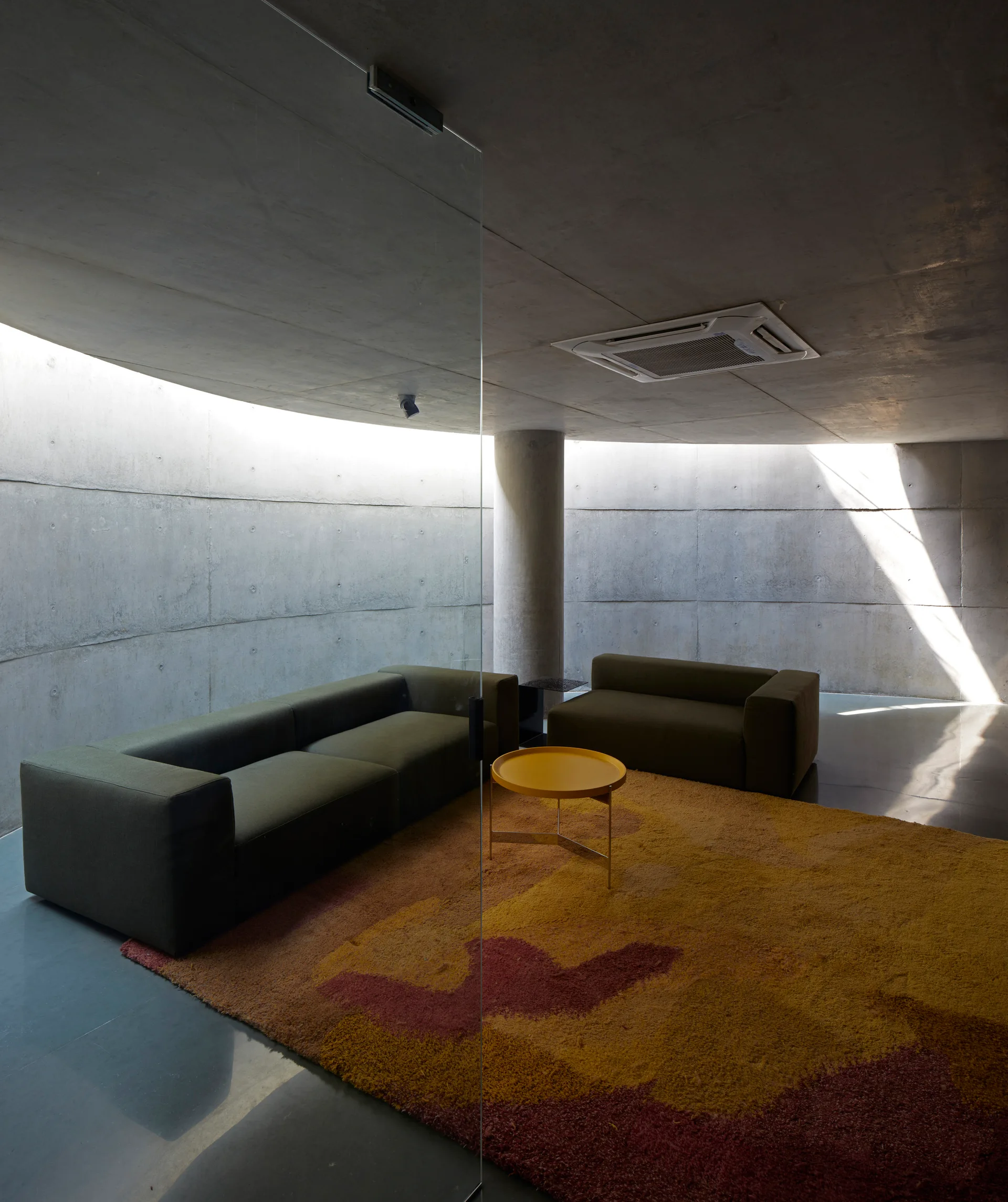 Olive green sofas sit on a warm yellow and red textured rug in a minimalist living room with curved concrete walls and a glass partition.