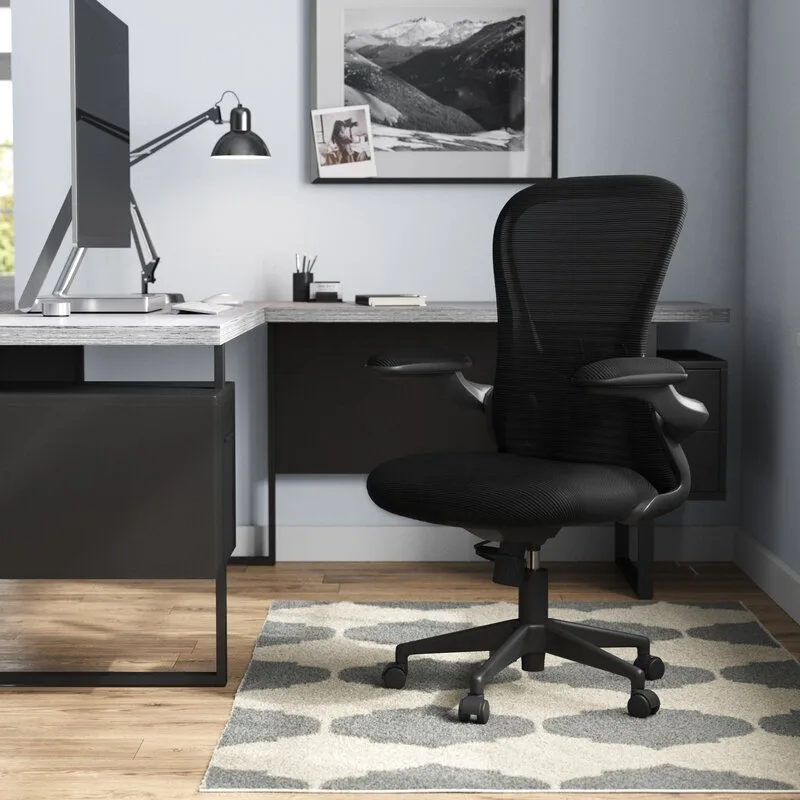 A black ergonomic mesh office chair sits on a grey patterned rug next to an L-shaped desk with a monitor.