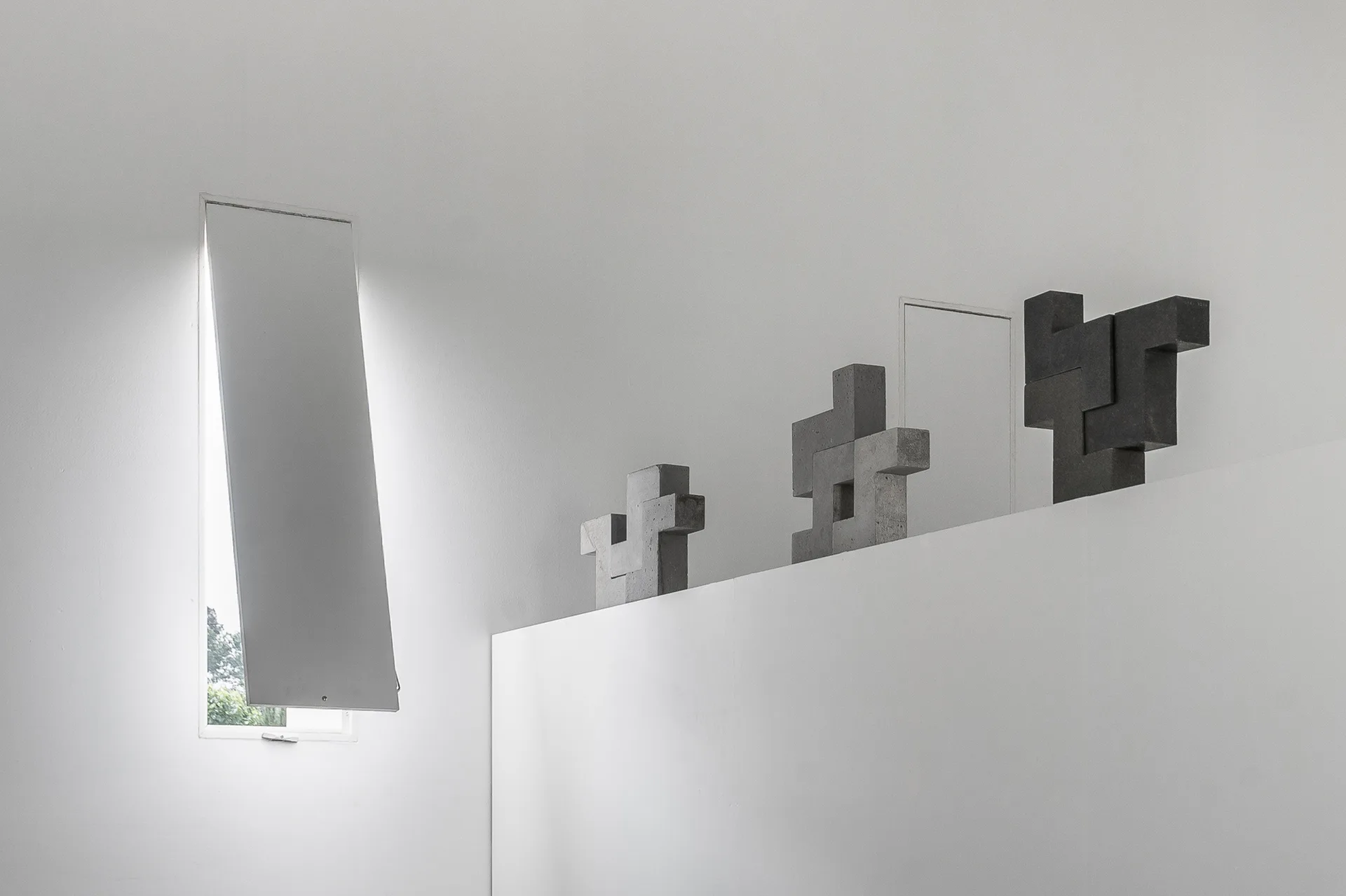 Three gray geometric stone sculptures stand on a high white ledge next to a narrow window with an open angled shutter.
