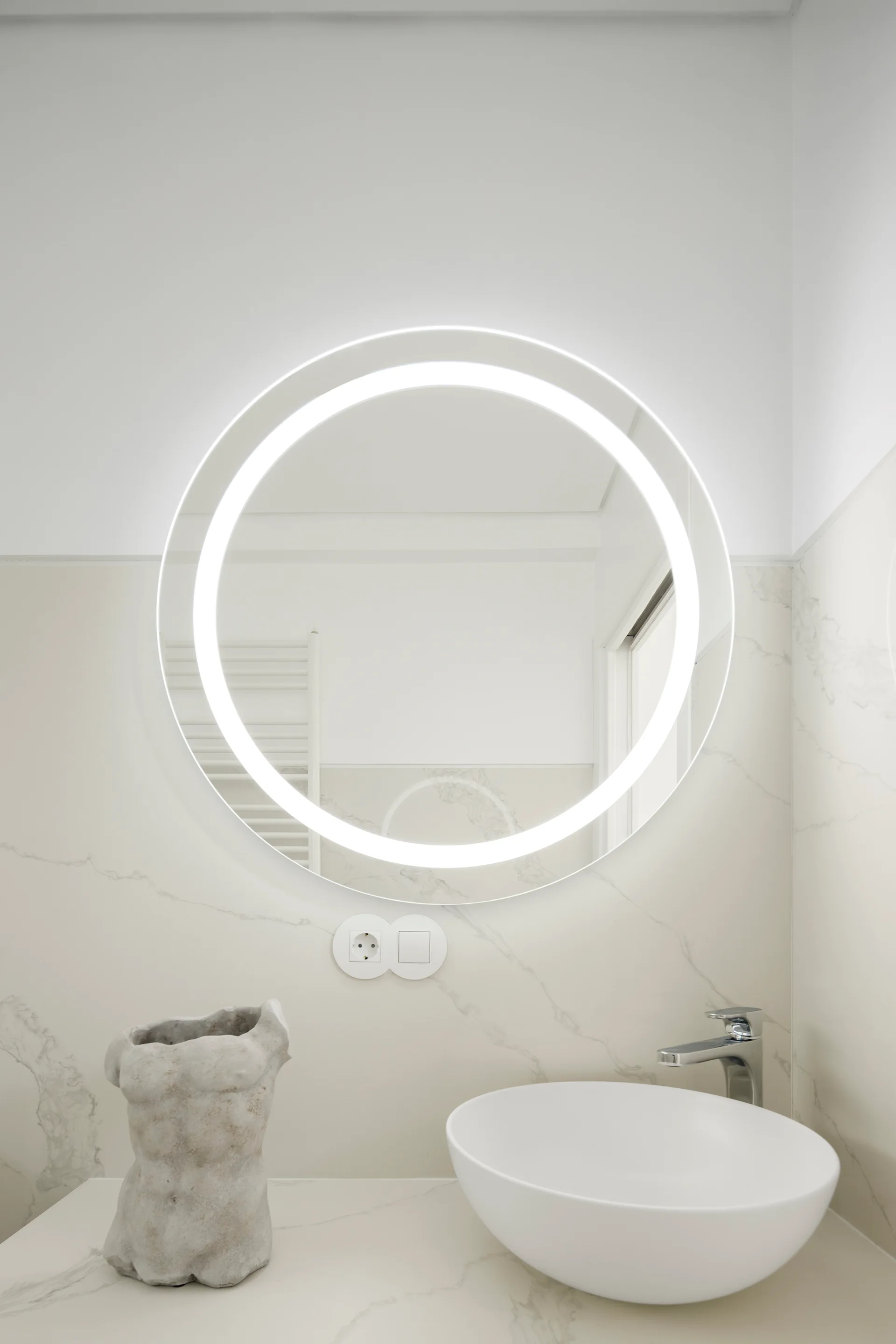 A minimalist bathroom vanity features a glowing circular backlit mirror on a veined marble tile wall above a white ceramic vessel sink.