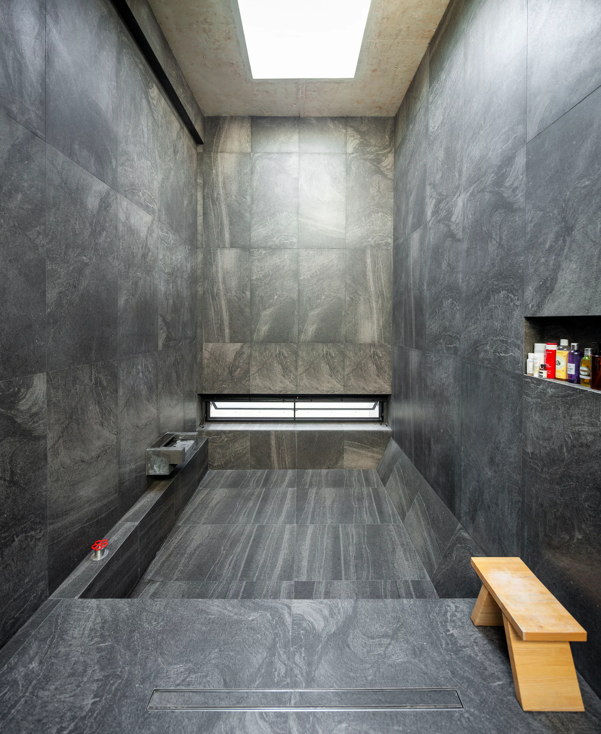 A minimalist walk-in shower room wrapped in charcoal grey stone tile features a large skylight and a small wooden bench.
