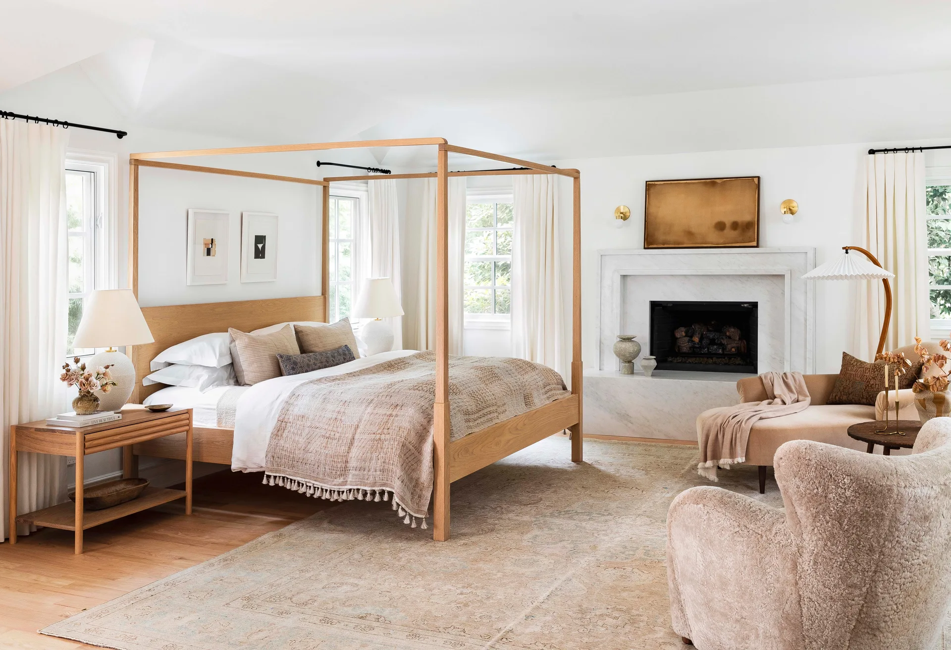 A bright bedroom features an oak canopy bed, a white marble fireplace, and neutral upholstered seating on a patterned rug.