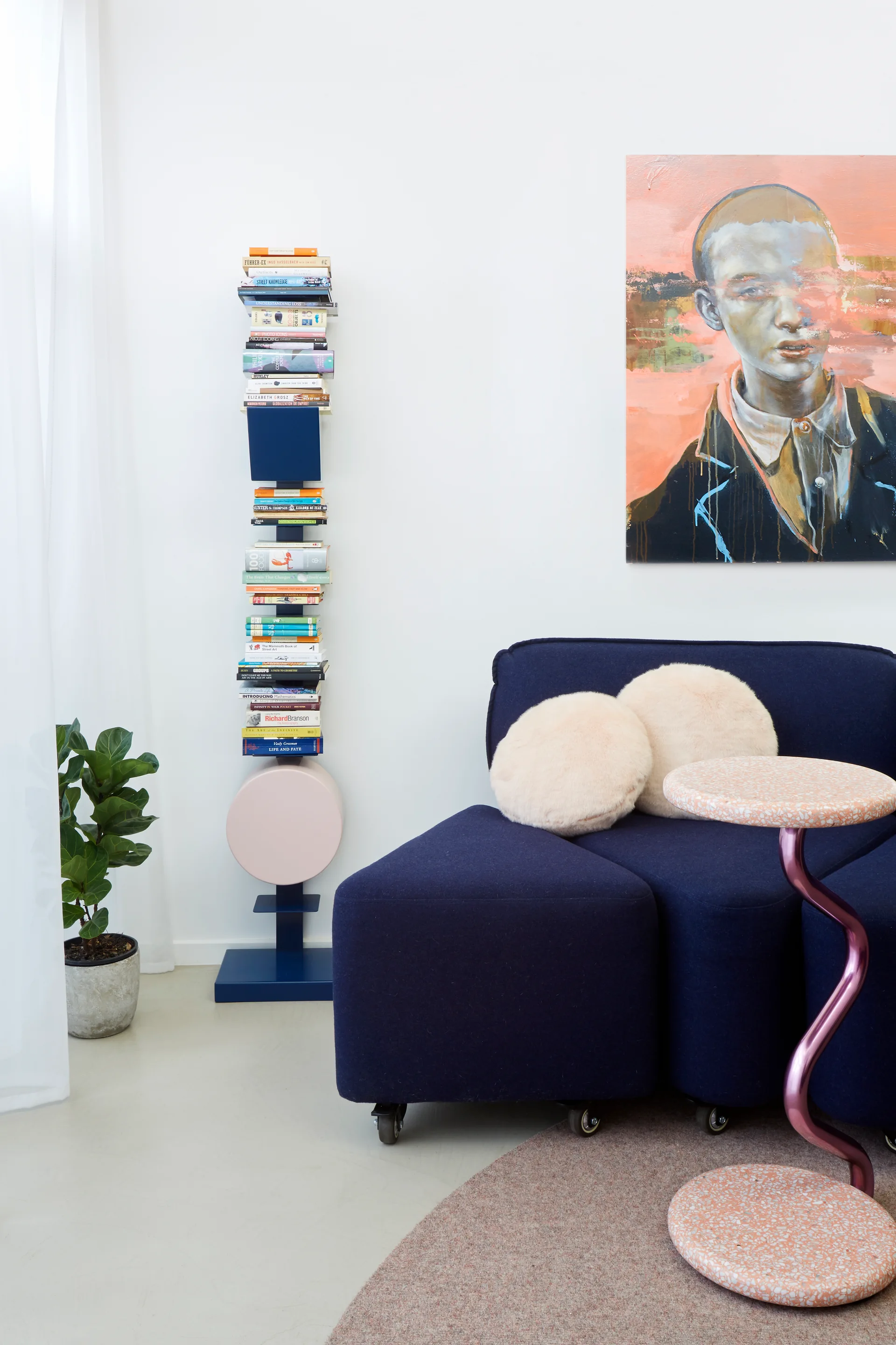 A navy modular sofa sits next to a tall vertical spine bookshelf stacked with books under a large portrait painting on a white wall.