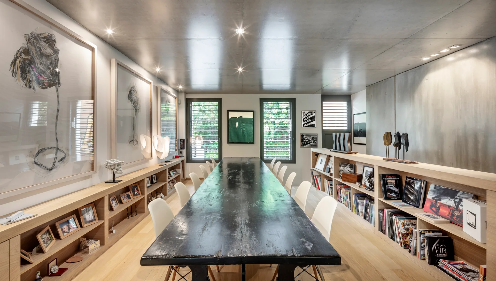A spacious dining room features a long dark wood table surrounded by white chairs, flanked by low light-wood built-in bookshelves under a grey ceiling.