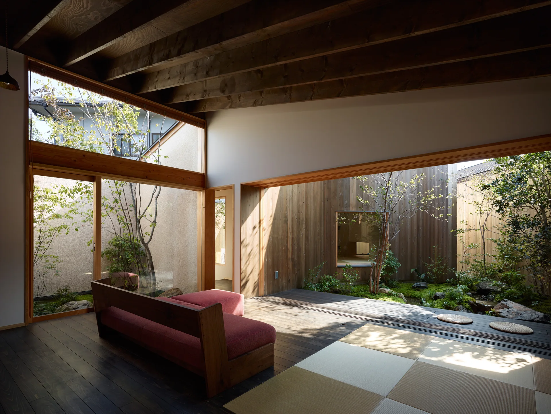 A minimalist room with a red sofa, tatami floor mats, and exposed ceiling beams next to large glass doors opening to a green courtyard garden.