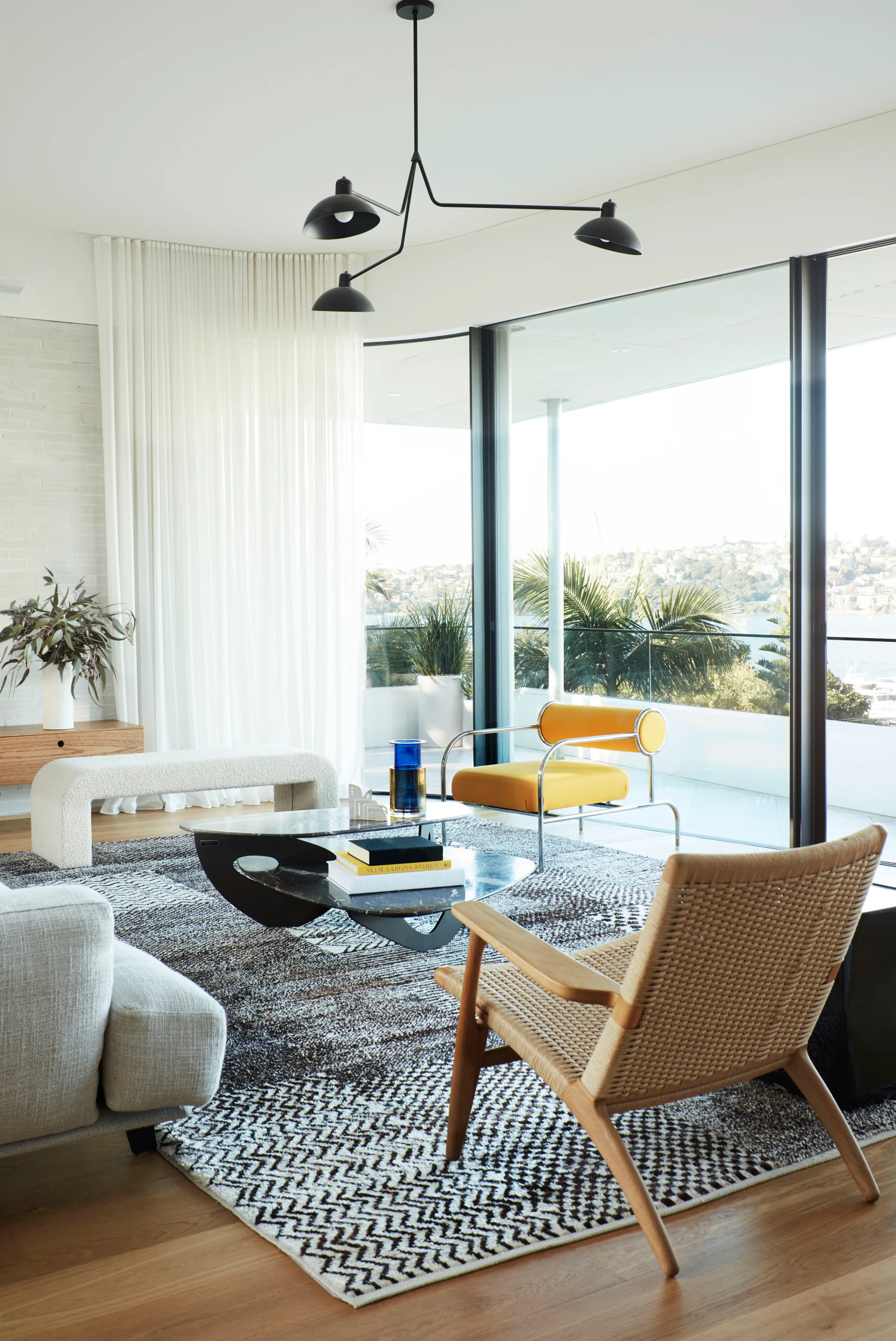 A modern living room features a yellow armchair, a woven wood chair, a black patterned rug, and floor-to-ceiling curved windows with a waterfront view.