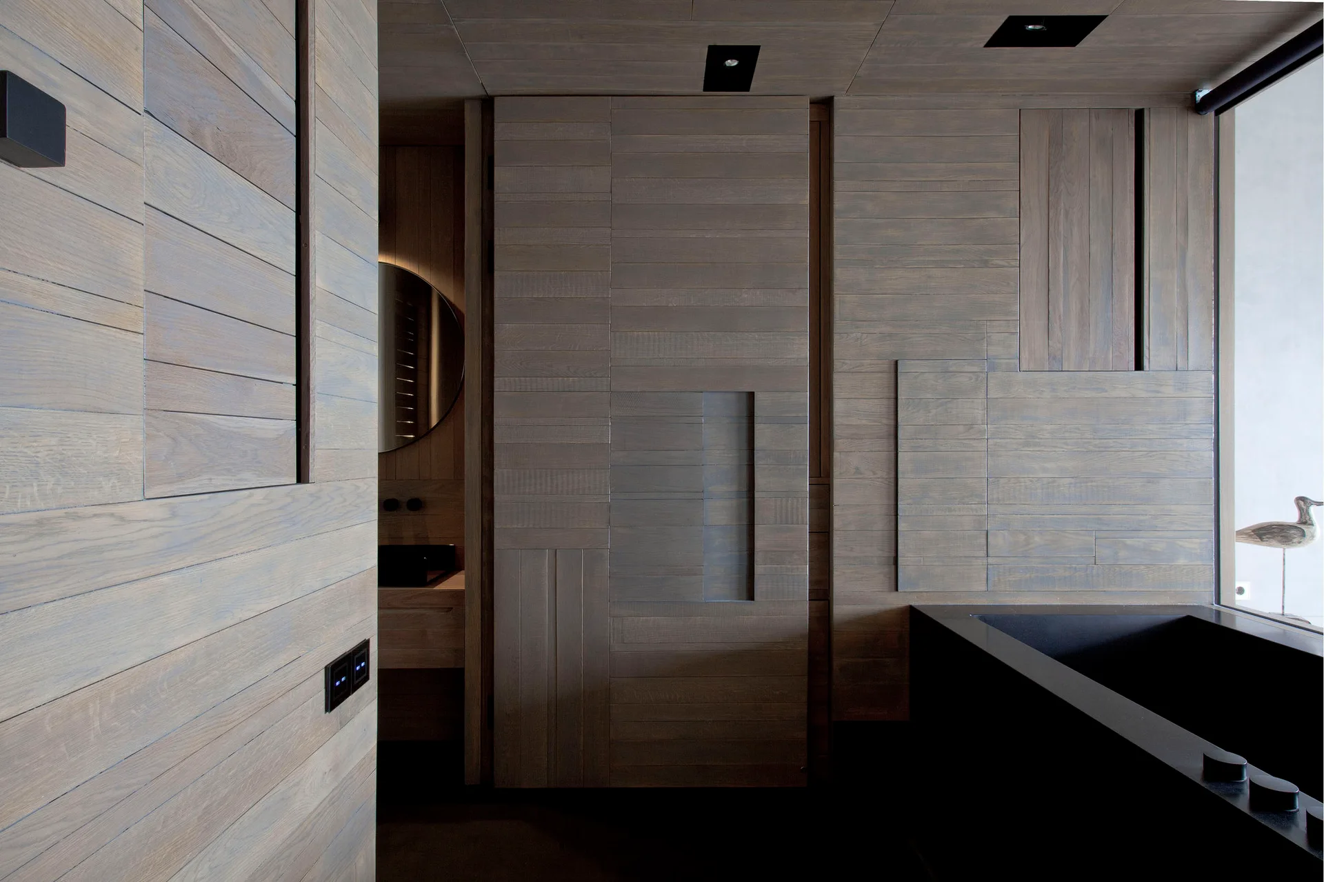 A minimalist bathroom with walls and ceiling clad in horizontal grey wood panels features a black rectangular bathtub.