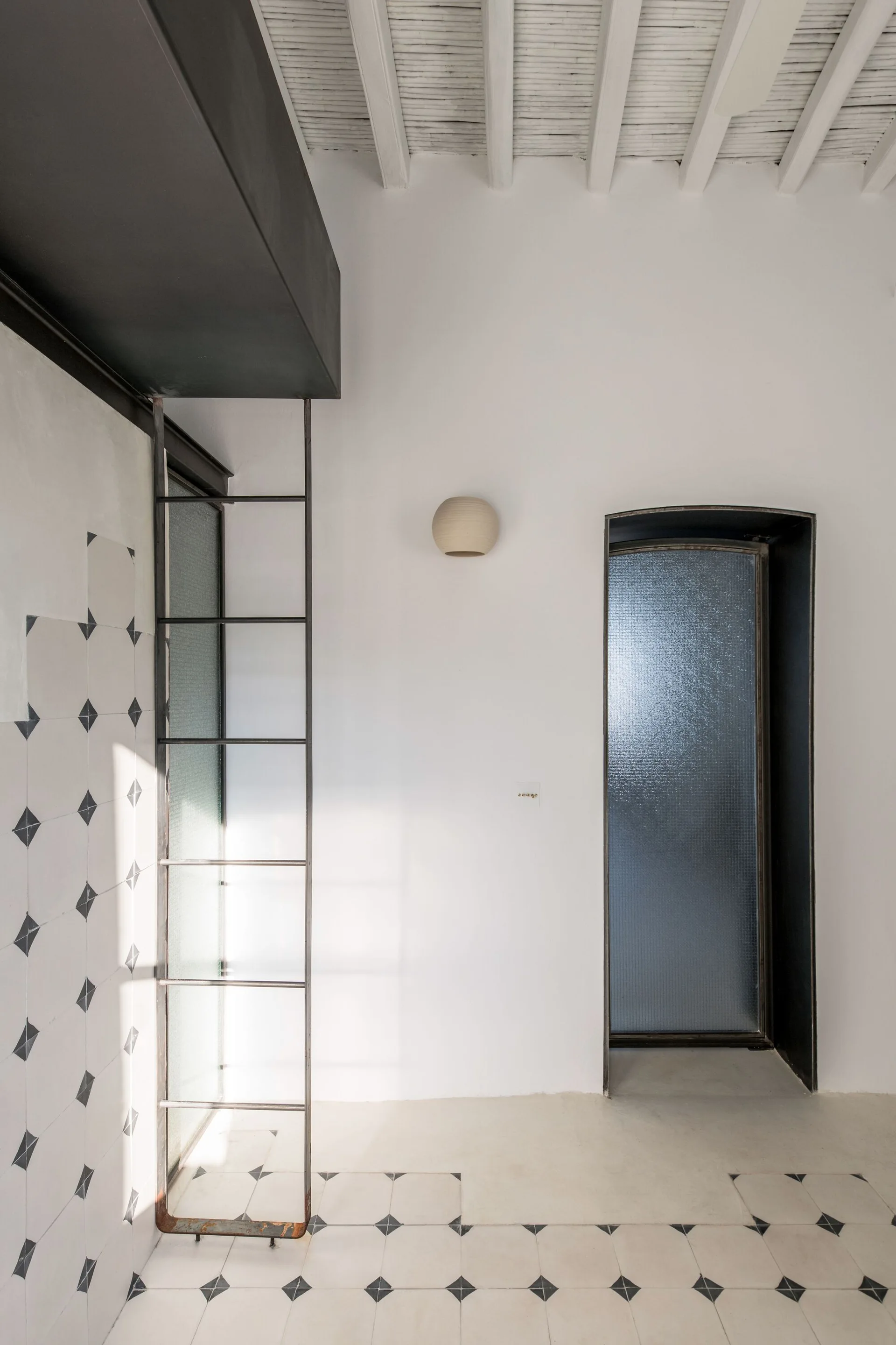 A tall dark metal ladder stands against a white-tiled wall with a black diamond pattern under a white beamed ceiling near an arched frosted glass door.