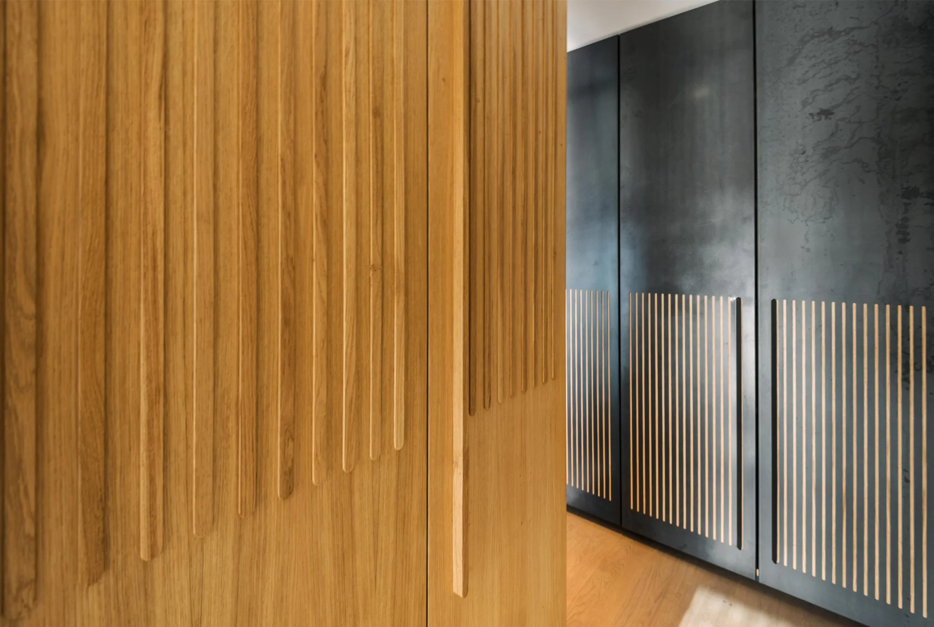 Light wood cabinets with vertical slats stand near dark charcoal closet doors featuring matching slatted cutouts.