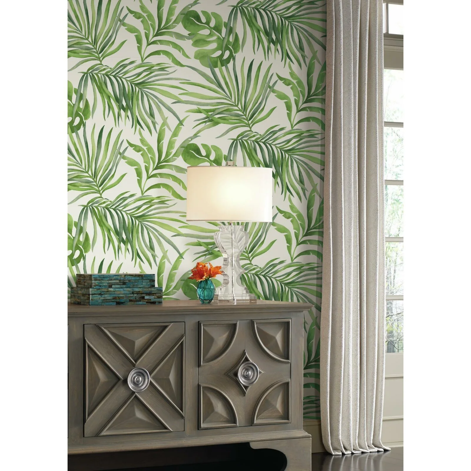 A wooden console cabinet with geometric door panels stands against a wall with green tropical leaf wallpaper next to a cream curtain.