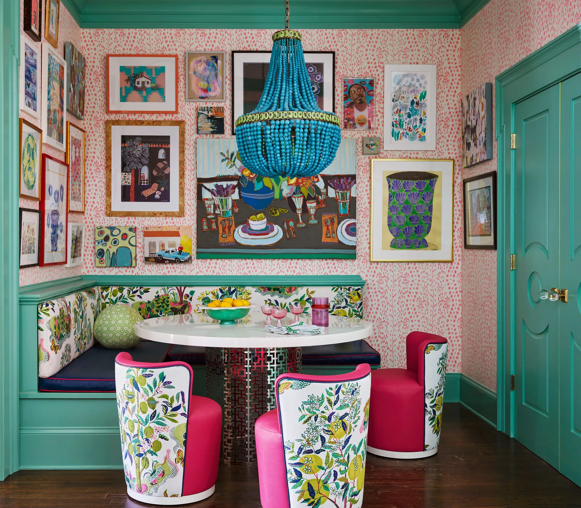 A colorful dining nook with a white table, teal banquette, pink floral chairs, a blue beaded chandelier, and a gallery wall.
