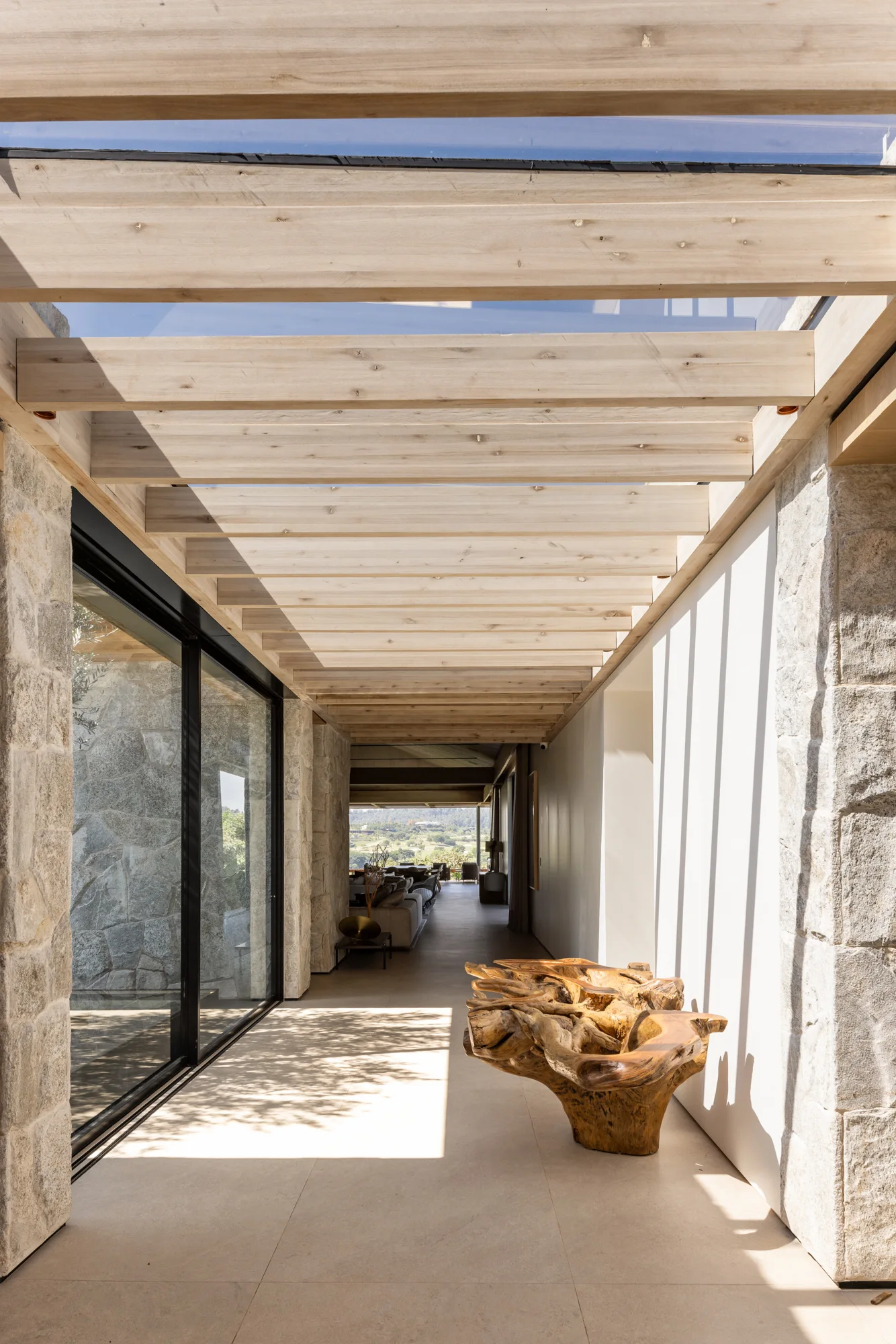 A sunlit hallway features exposed wood ceiling beams, a rugged stone wall, and a large natural tree root bench on a tiled floor.