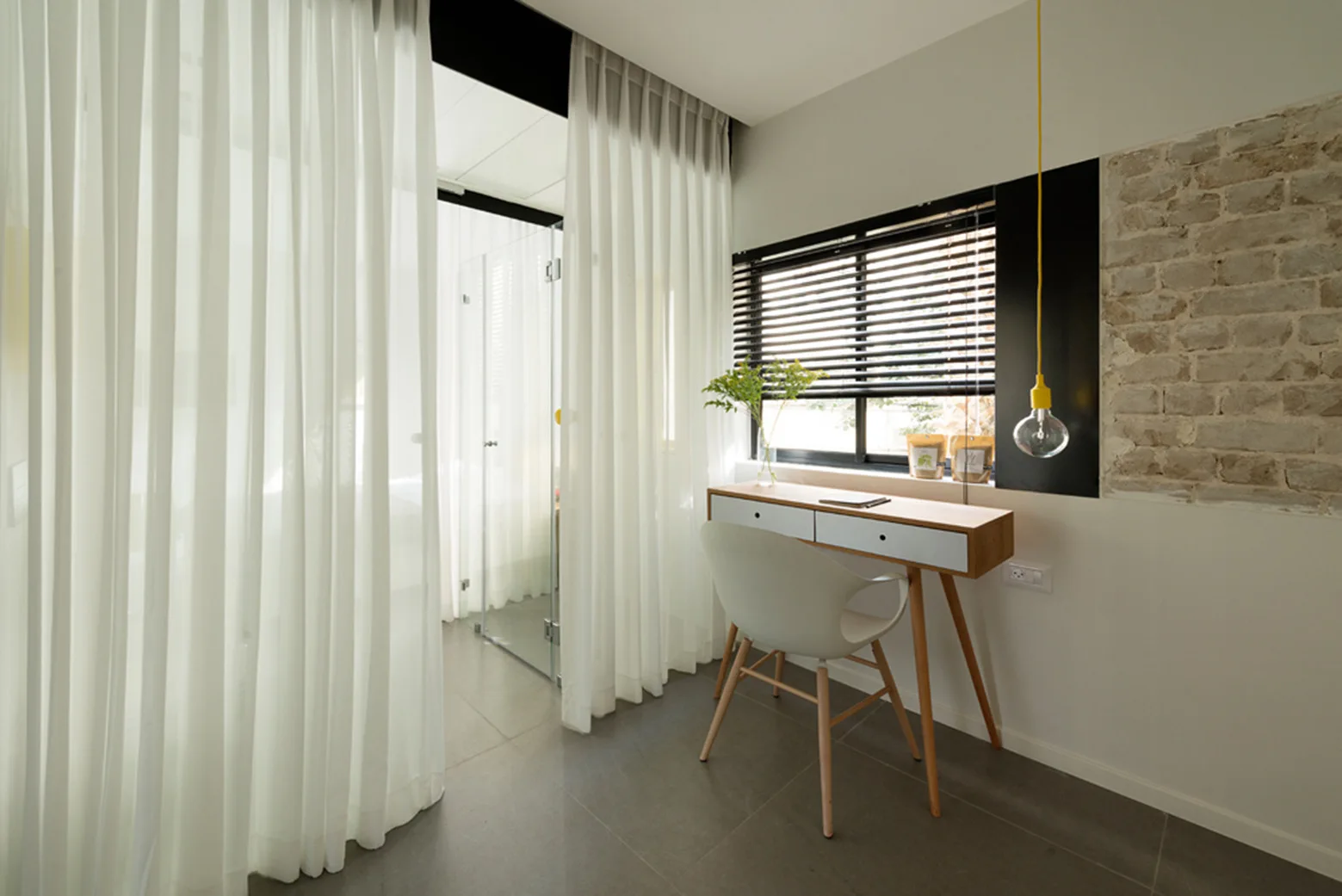 A minimalist home office featuring a light wood desk and white chair next to sheer white curtains, black window blinds, and an exposed brick wall section.