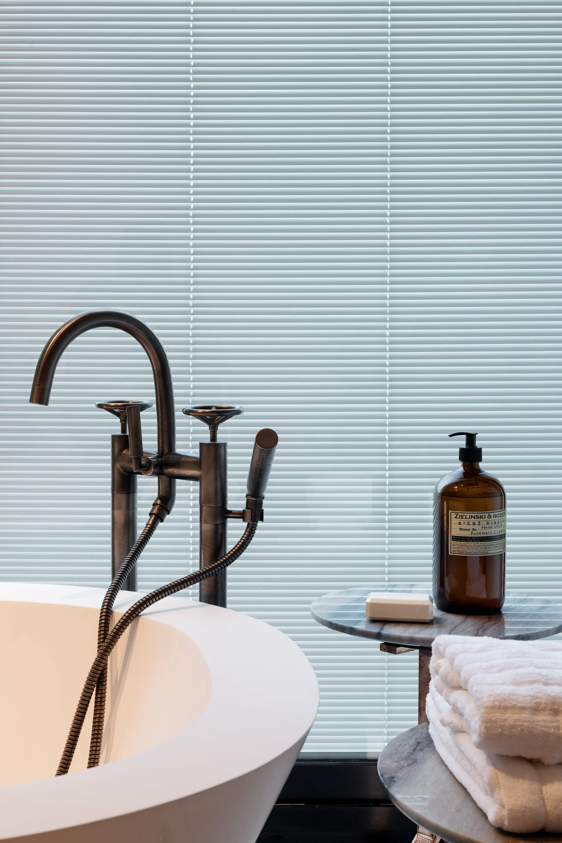 A close-up of a white freestanding bathtub with a dark bronze faucet, next to a tiered marble side table holding white towels and an amber bottle.