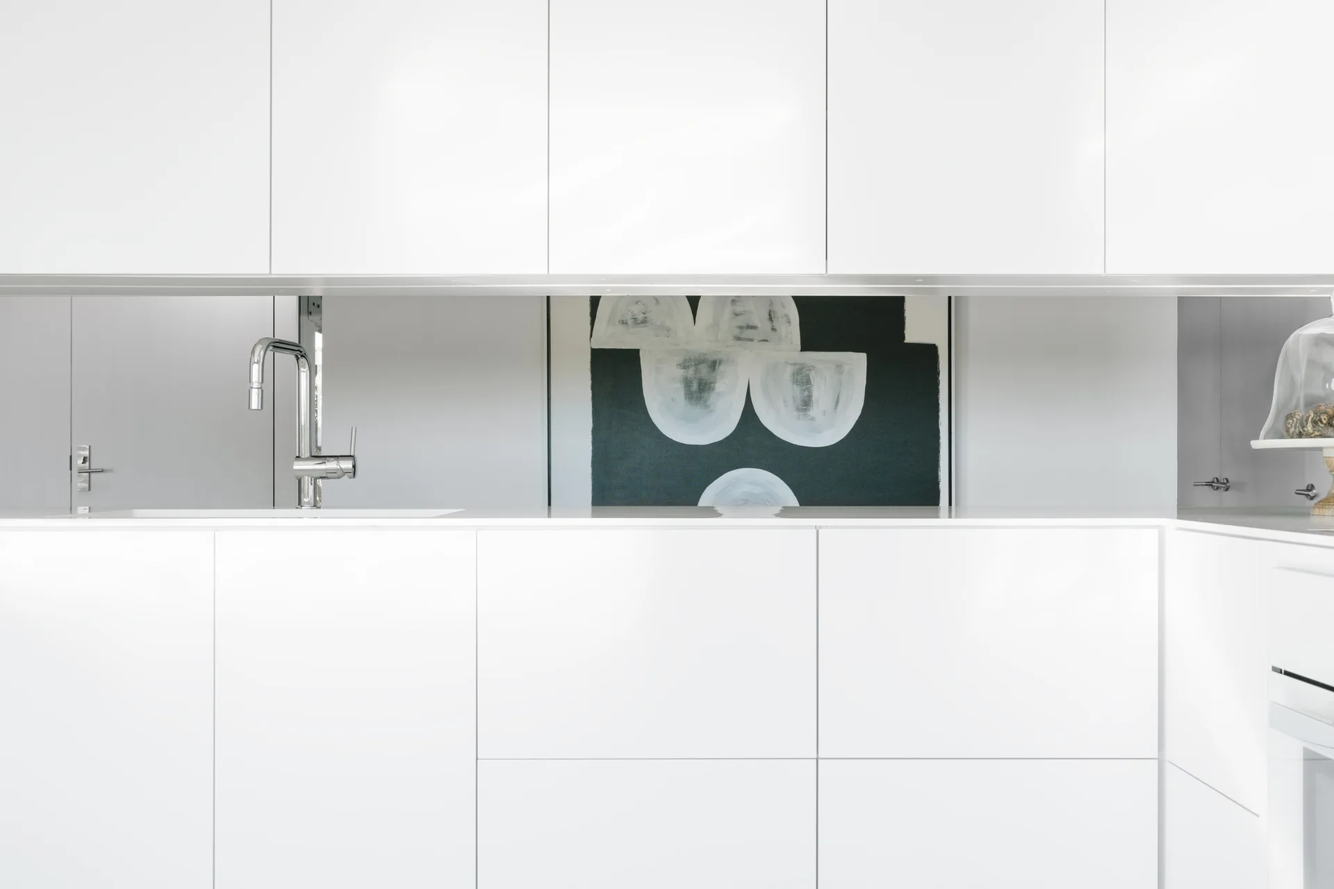 Sleek white flat-panel upper and lower kitchen cabinets surround a white countertop with a chrome faucet and a framed black and white abstract painting.