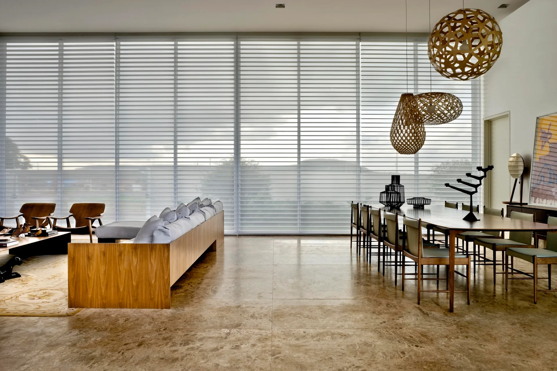 An open-concept living and dining area features a large wood-framed sofa, a long wooden dining table, stone floors, and woven sphere pendant lights.