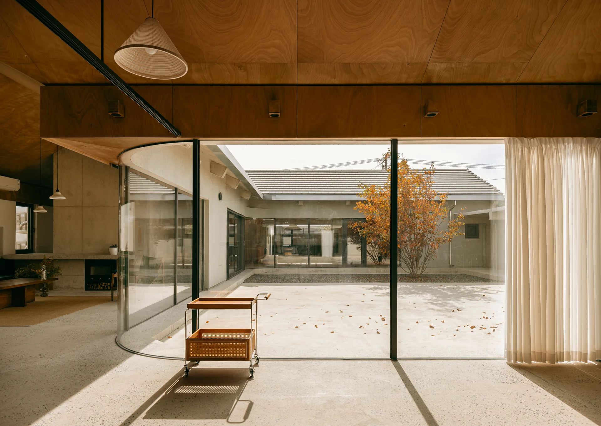 A minimalist room with a curved glass facade facing an outdoor courtyard, featuring a wood paneled ceiling and a small wood serving cart.