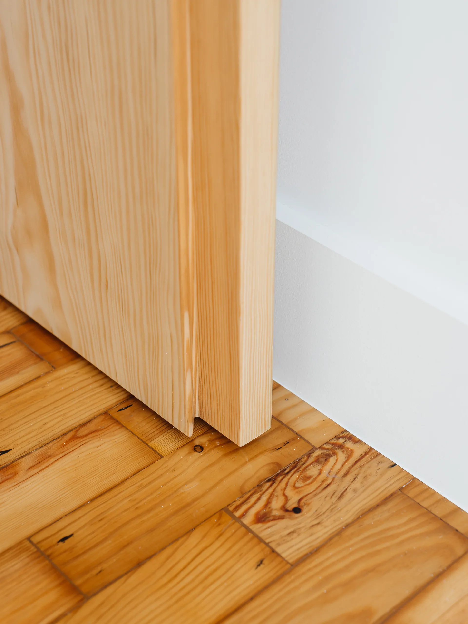 A close-up of a light-colored wooden door frame meeting a smooth white wall with a white baseboard on a wood plank floor.