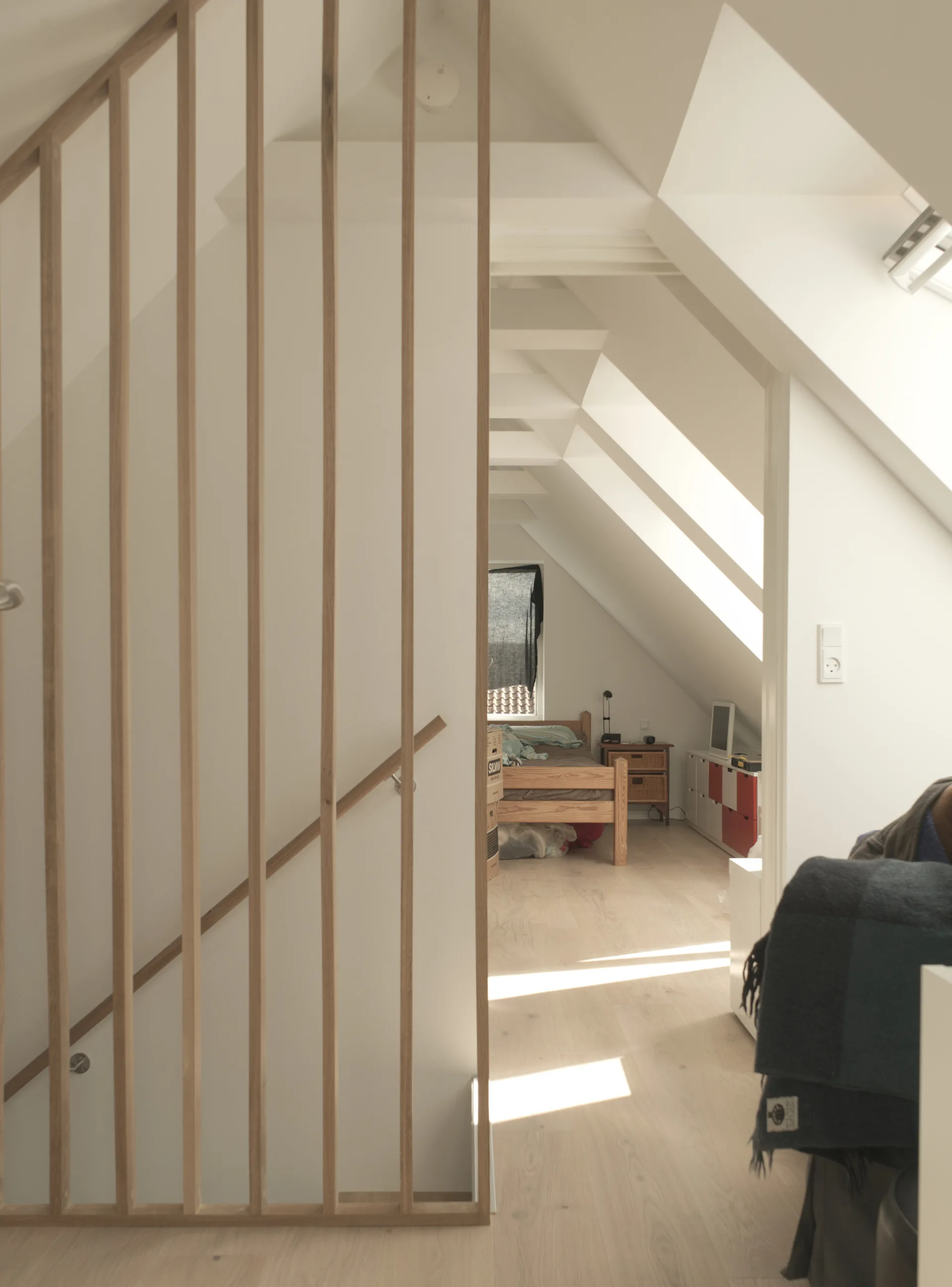 A view from a light wood staircase landing through vertical slats into a bright attic bedroom with sloped ceilings and skylights.