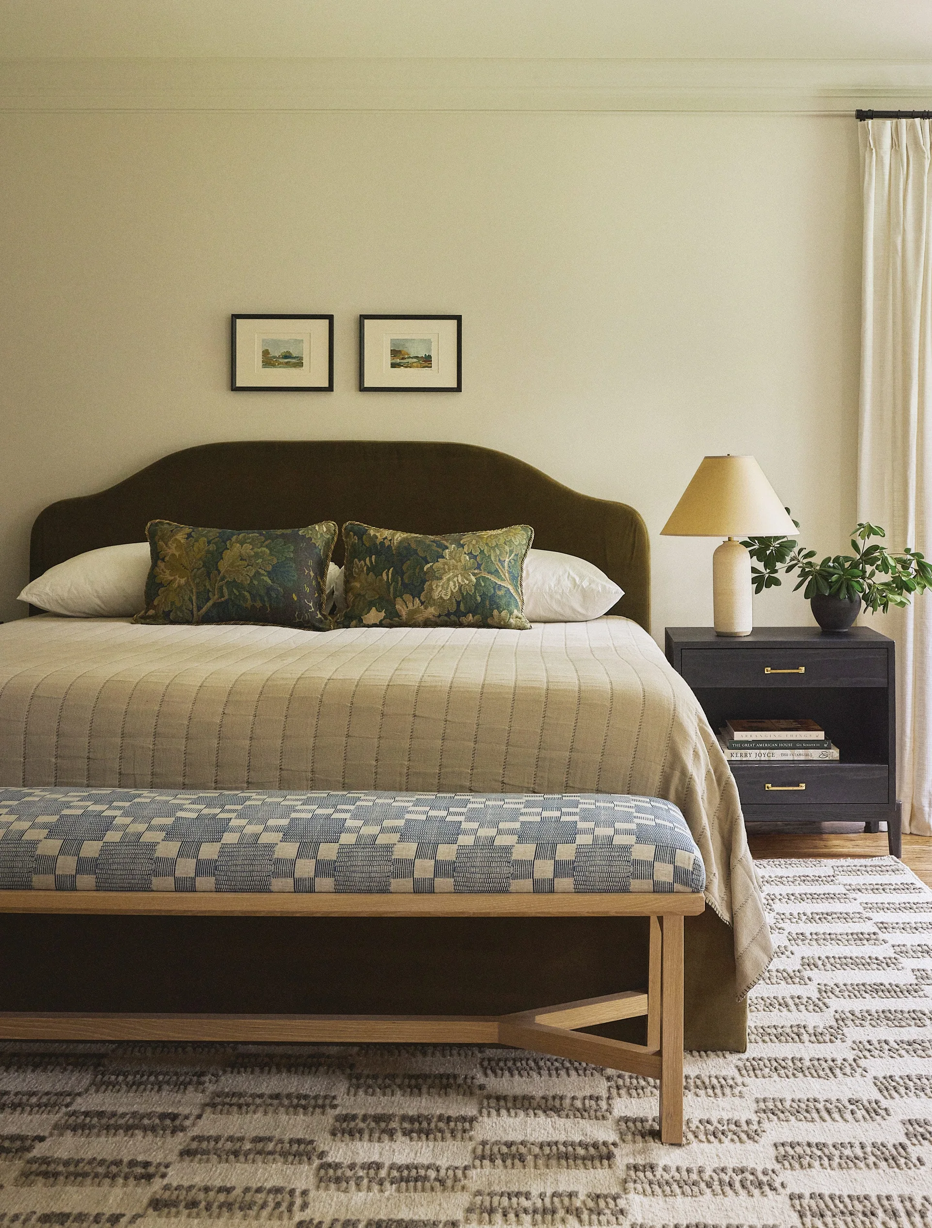 A bedroom features an olive green curved headboard, a dark nightstand with a lamp, and a patterned blue bench at the foot of the bed.