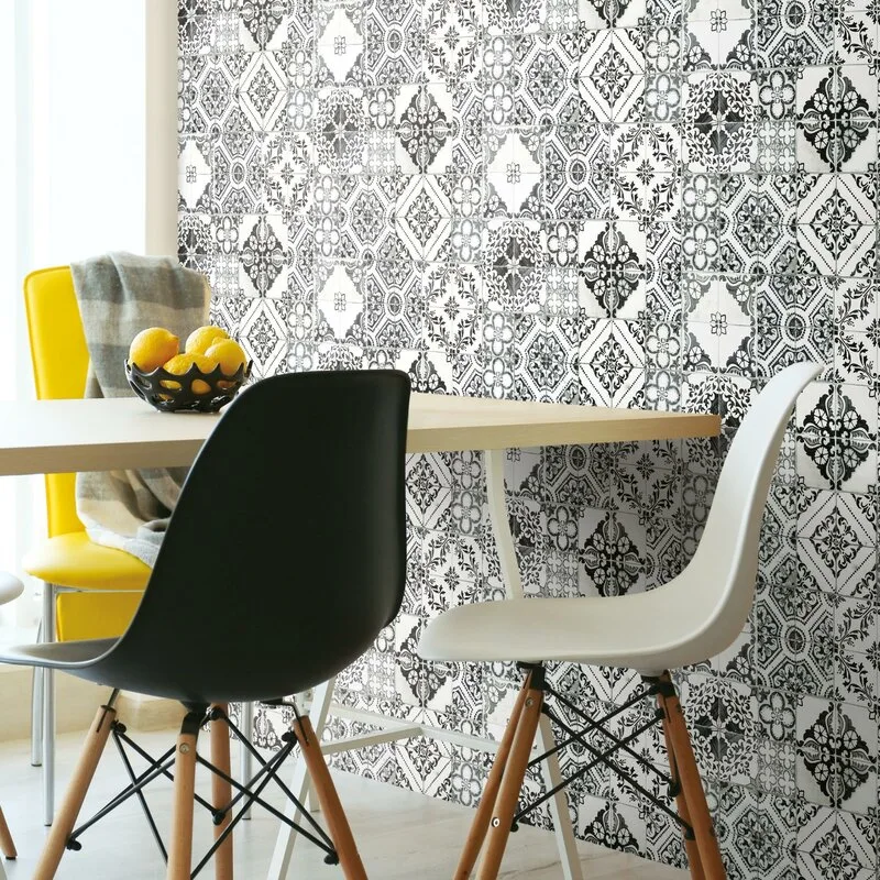 A light wood dining table with black, white, and yellow chairs set against a detailed black and white patterned tile accent wall.