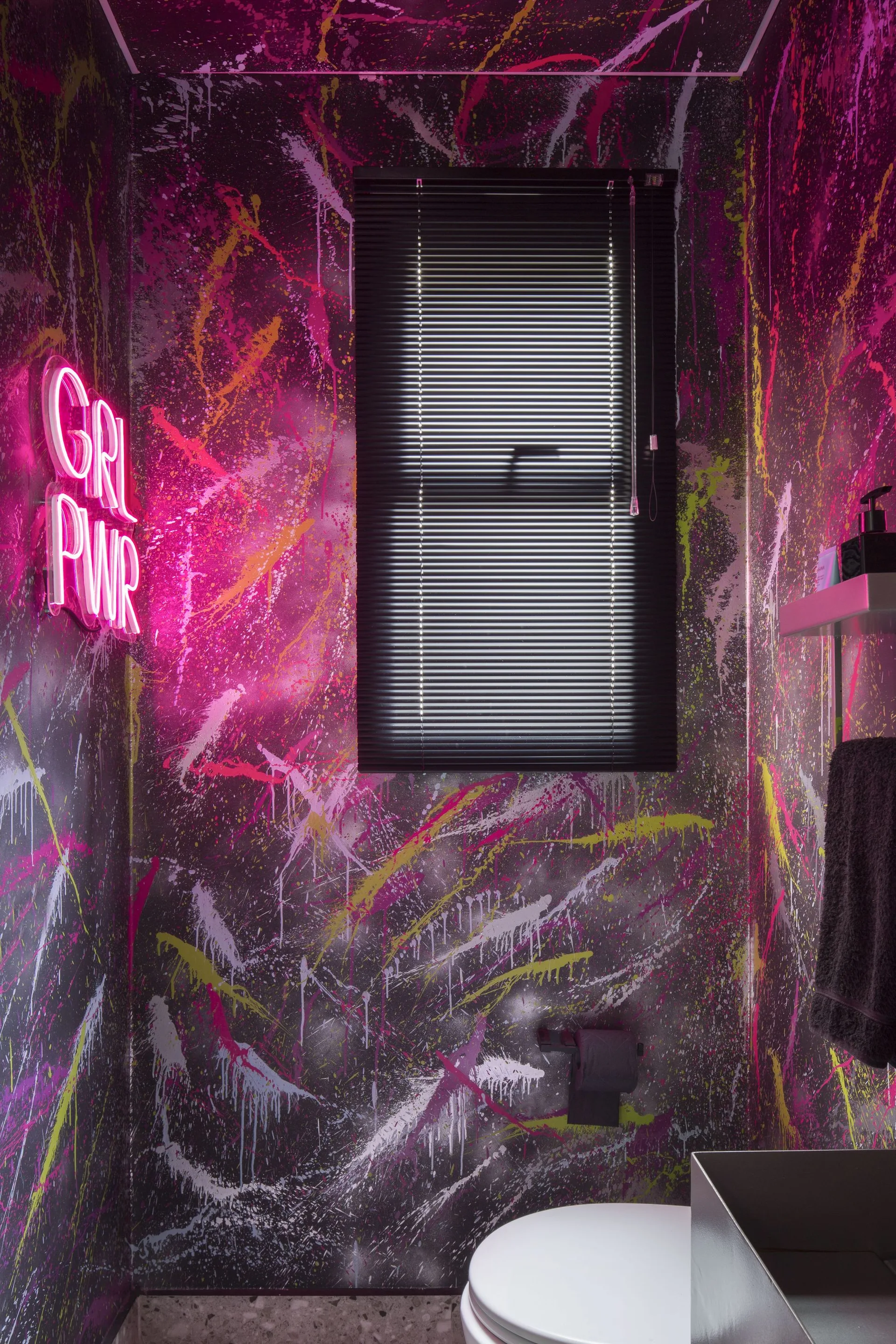 A small powder room featuring black walls and ceiling covered in multi-colored paint splatters, a glowing pink neon sign, and black window blinds.
