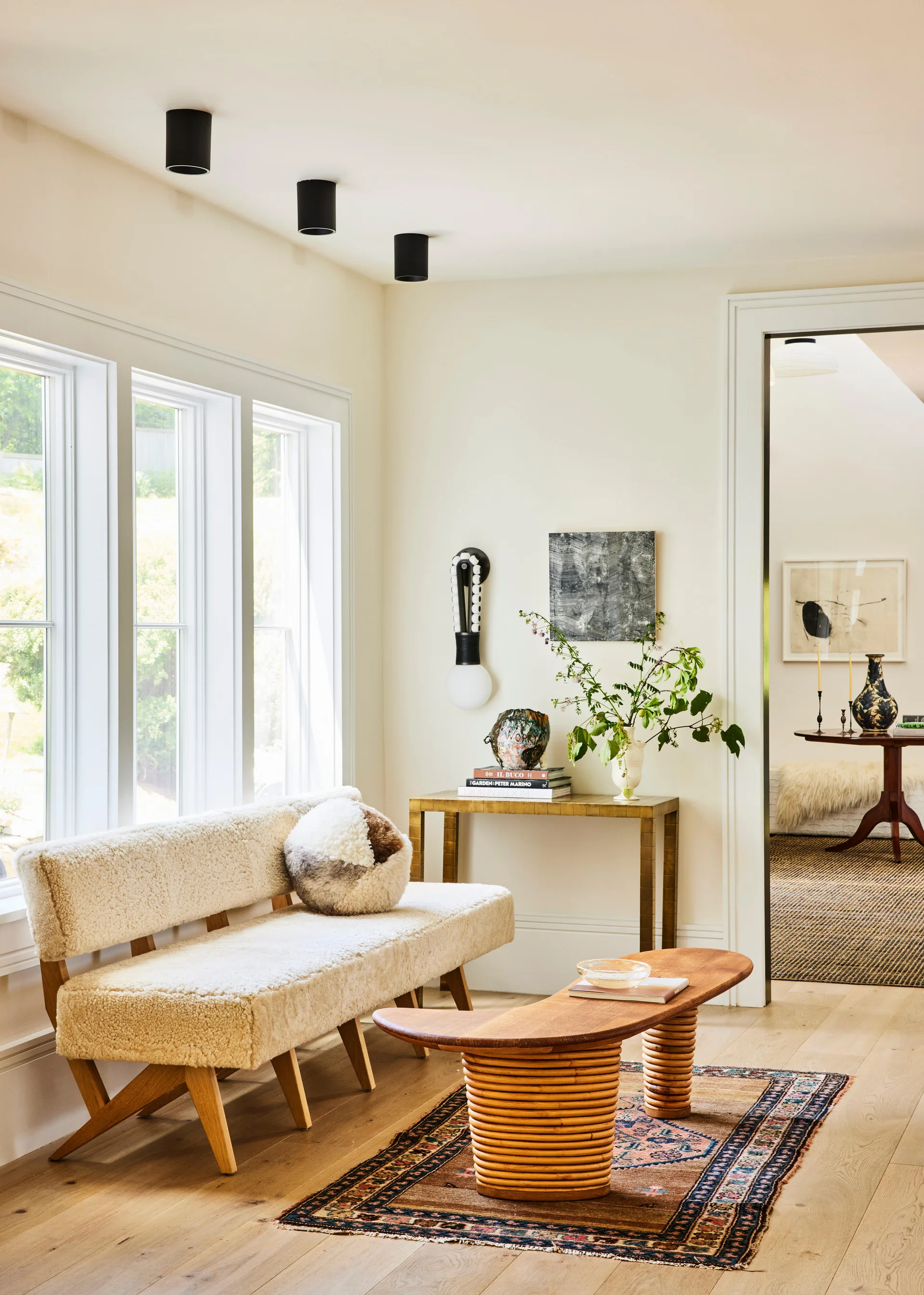 A bright living space features a sheepskin upholstered bench, a curved wooden coffee table with a ribbed base, and an oriental rug on light wood floors.
