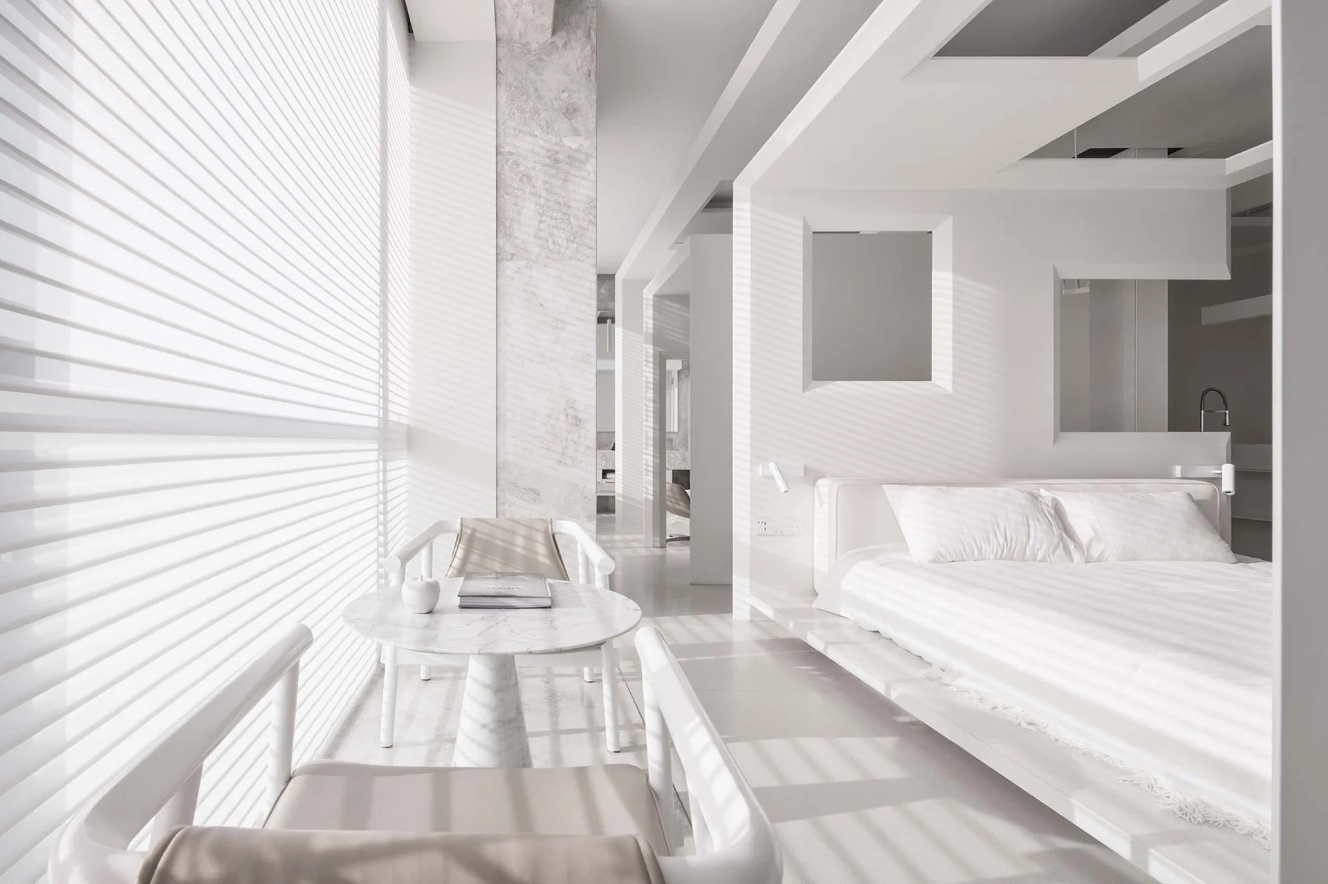 A minimalist white bedroom features a low platform bed and a small marble table with two armchairs near a window with horizontal blinds.