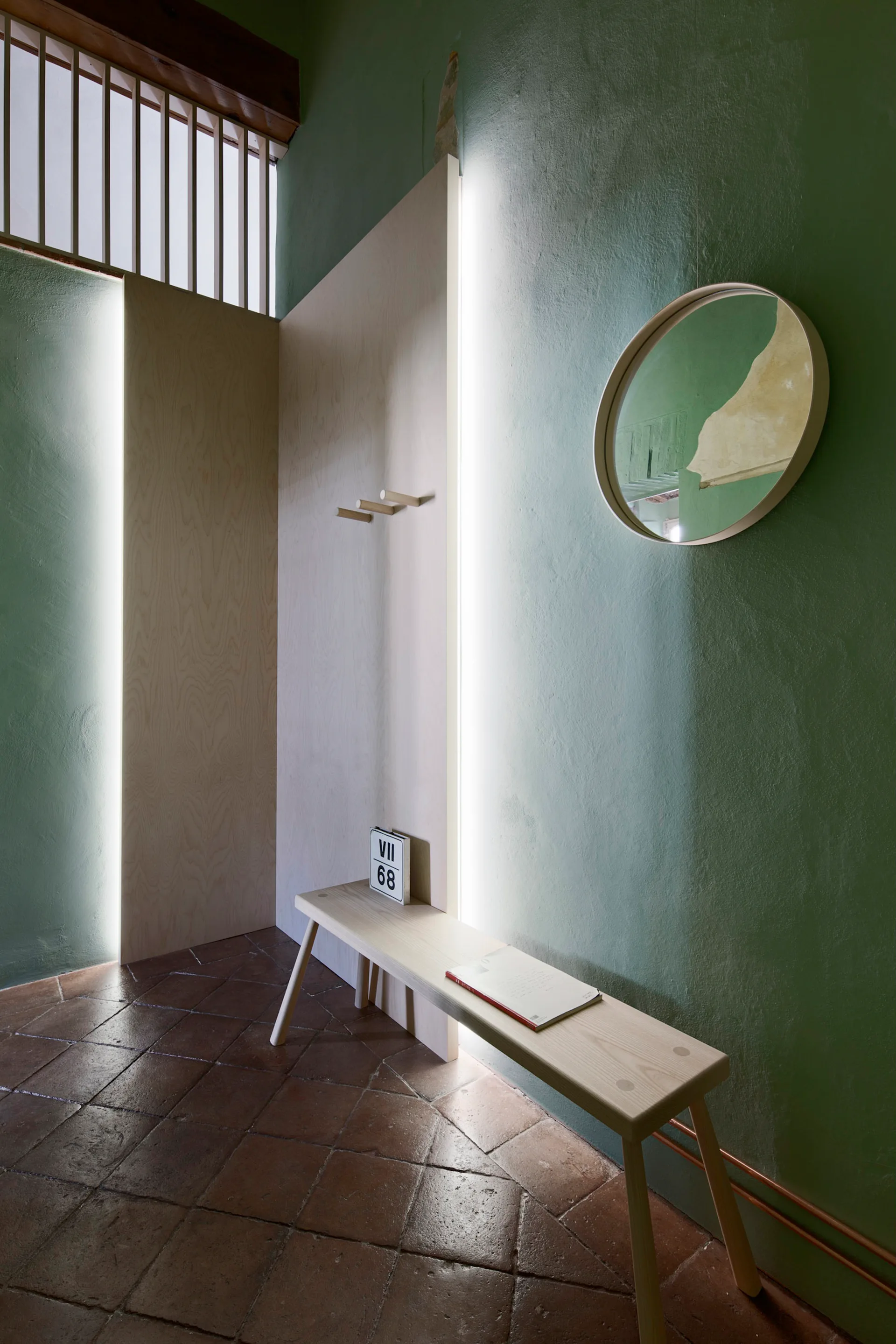 A minimalist entryway features a light wood bench, vertical wall panels with built-in LED lighting, green walls, and a terracotta tile floor.