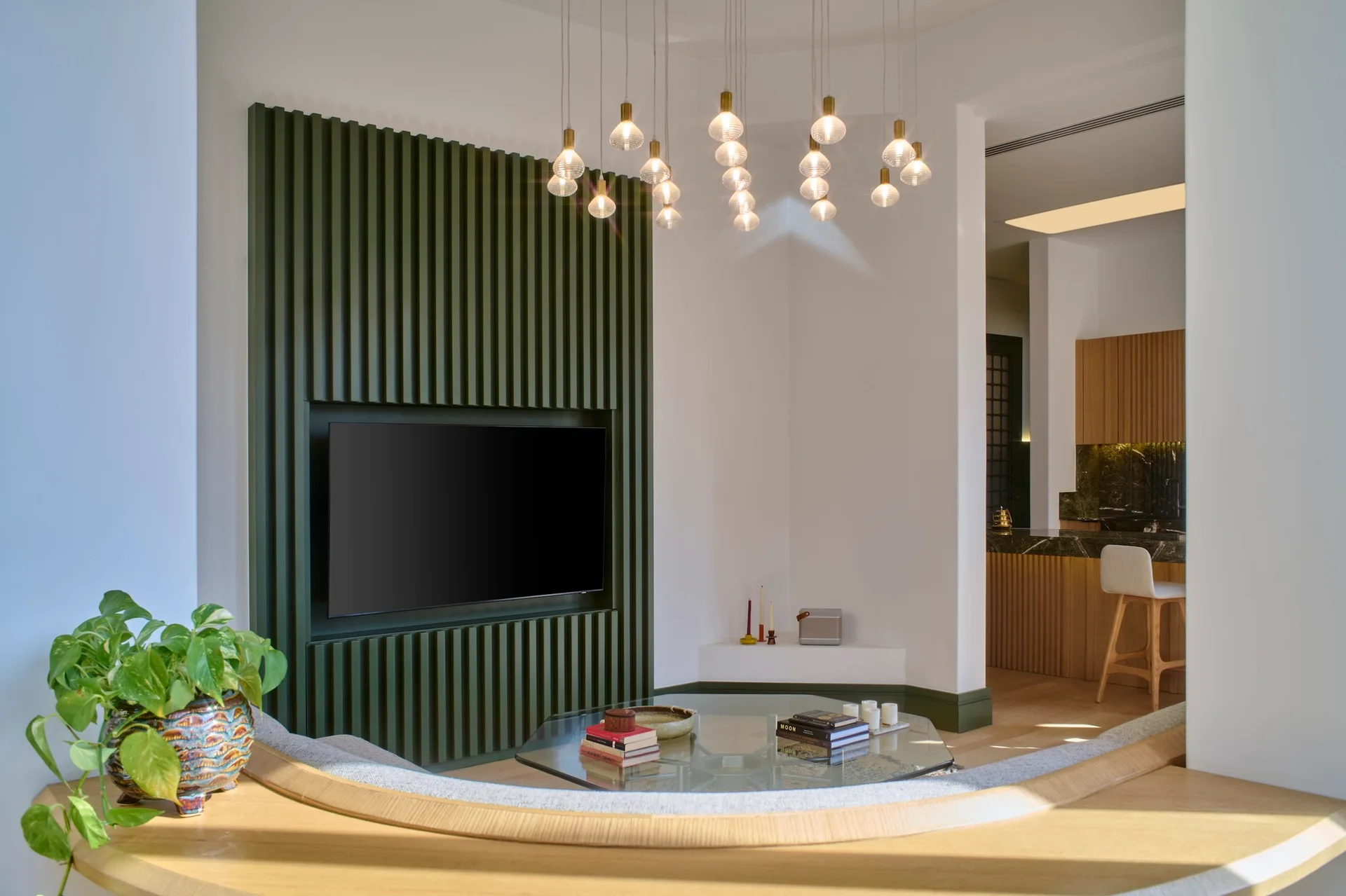 A modern living space with an olive green slatted media wall housing a TV, a hexagonal glass coffee table, and a cluster pendant light fixture.