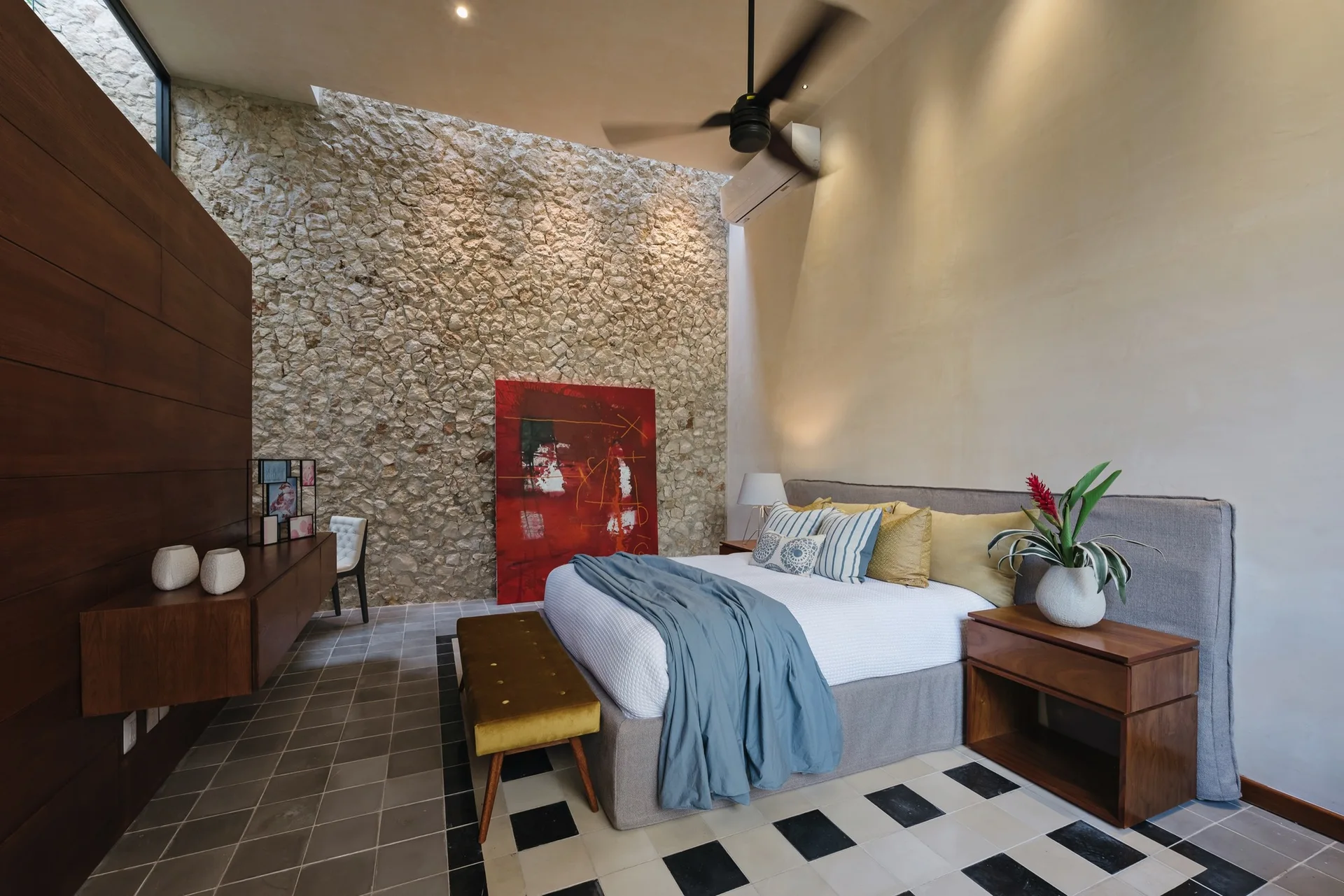 A modern bedroom with an upholstered bed, a textured stone accent wall beneath a skylight, a floating wood console, and checkered floor tile borders.