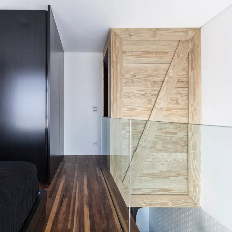 A contemporary landing featuring a frameless glass safety railing, dark hardwood floors, a matte black cabinet, and a light wood-clad staircase structure.