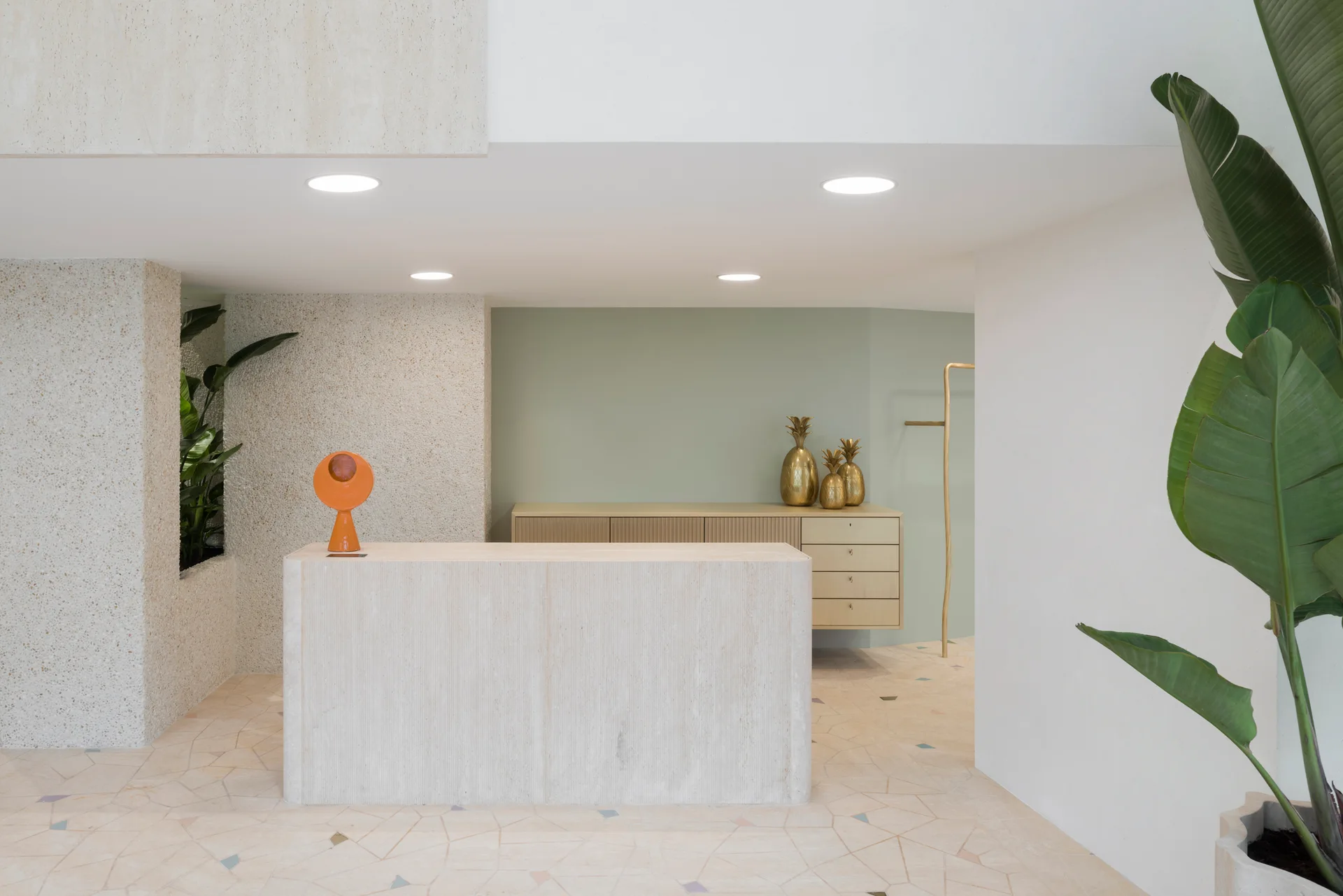 A minimalist reception area features a travertine stone desk, a sage green wall, a fluted wood cabinet, and speckled terrazzo flooring.
