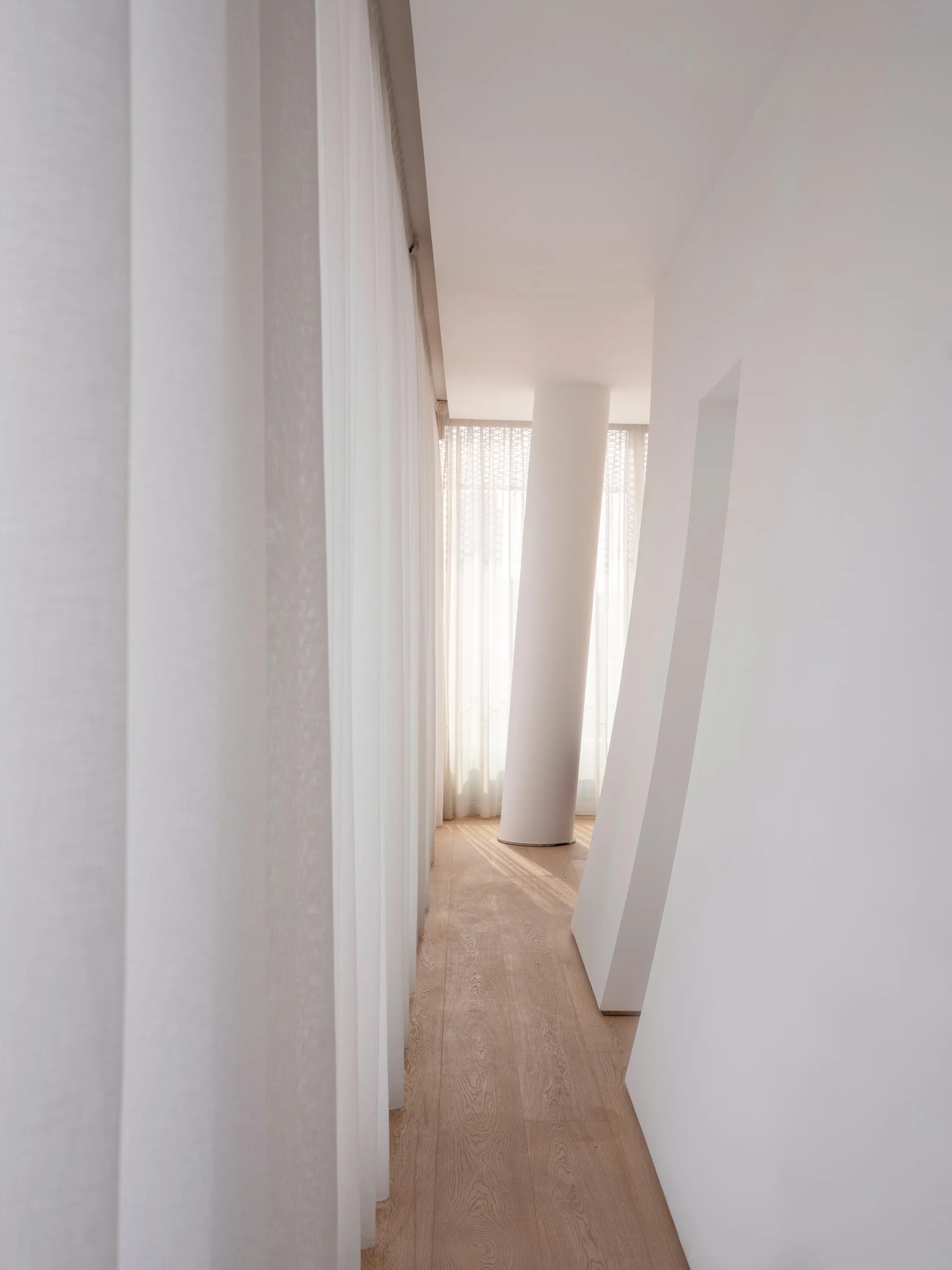 A bright minimalist hallway features light wood flooring, white walls, full-length sheer curtains, and a cylindrical white structural column.