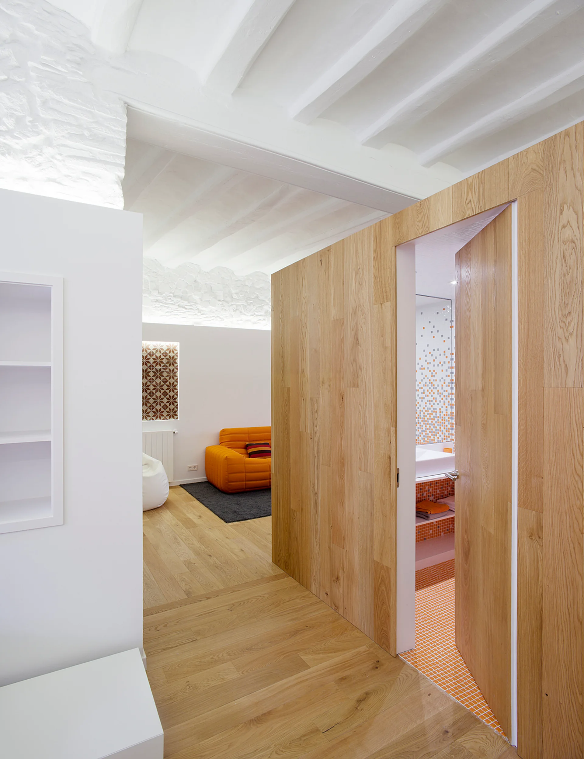 A light wood-paneled partition wall with an open door reveals a bathroom with orange mosaic tile next to a white living space with an orange sofa.