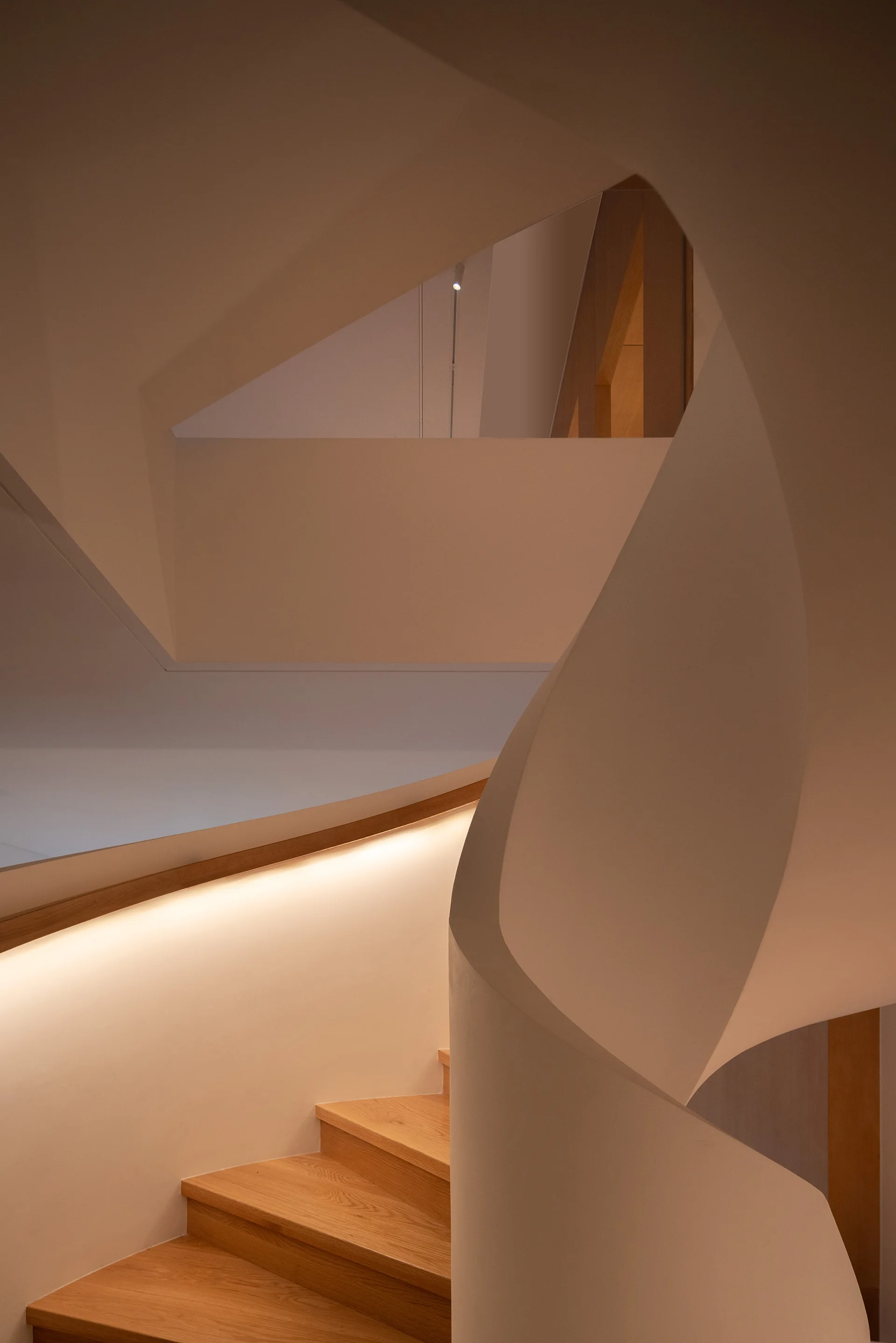 A modern spiral staircase features warm wood treads, a curved off-white plaster wall, and a wooden handrail with integrated warm strip lighting.