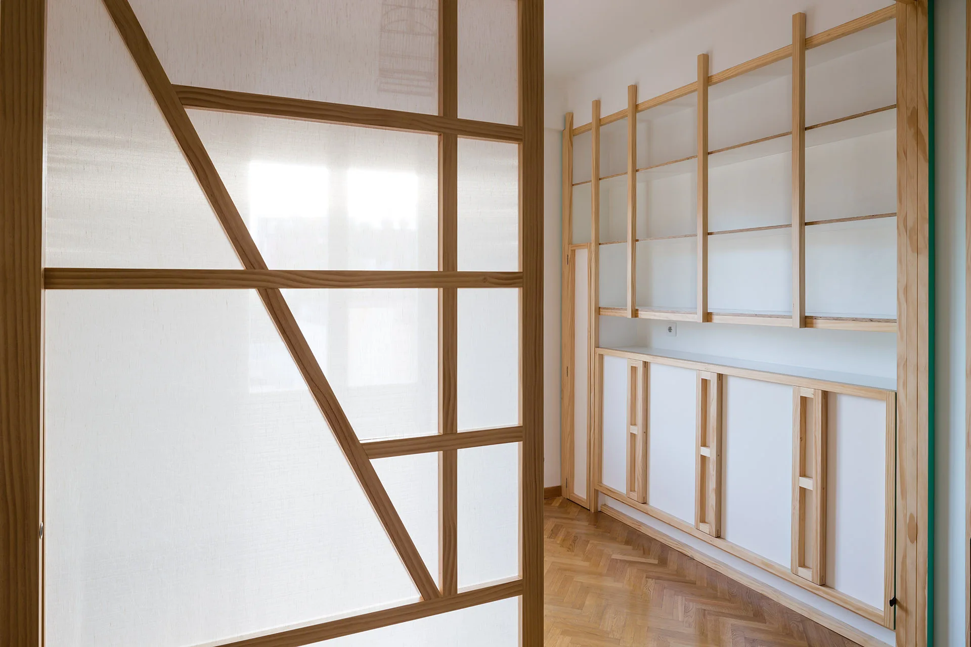 A light wood room divider with a fabric screen stands on a herringbone wood floor next to a matching built-in wall shelving unit.