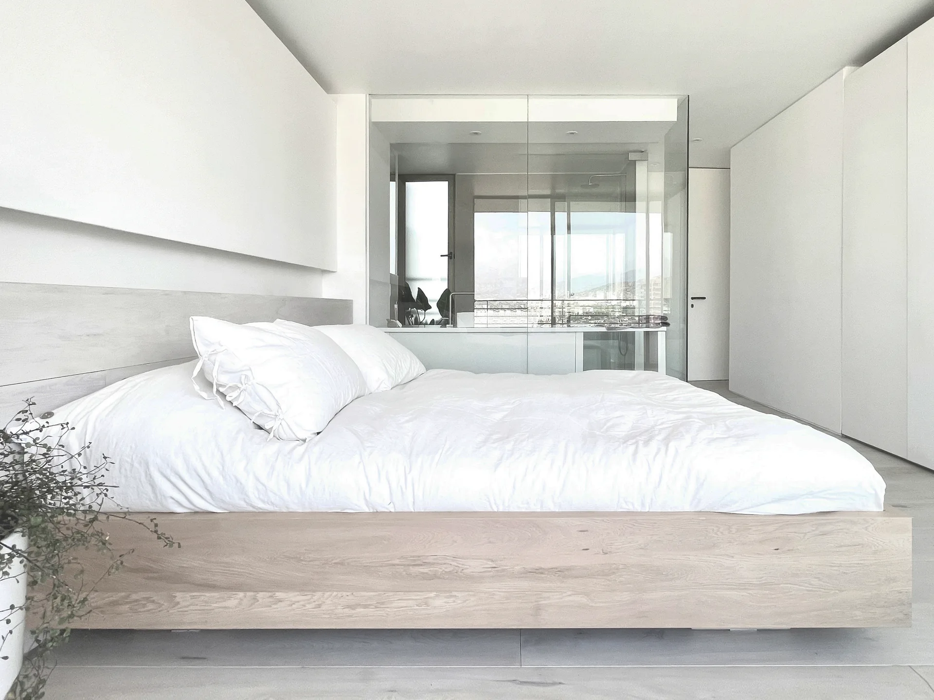 A minimalist bedroom features a low wooden platform bed with white bedding next to a floor-to-ceiling glass partition separating an ensuite bathroom.