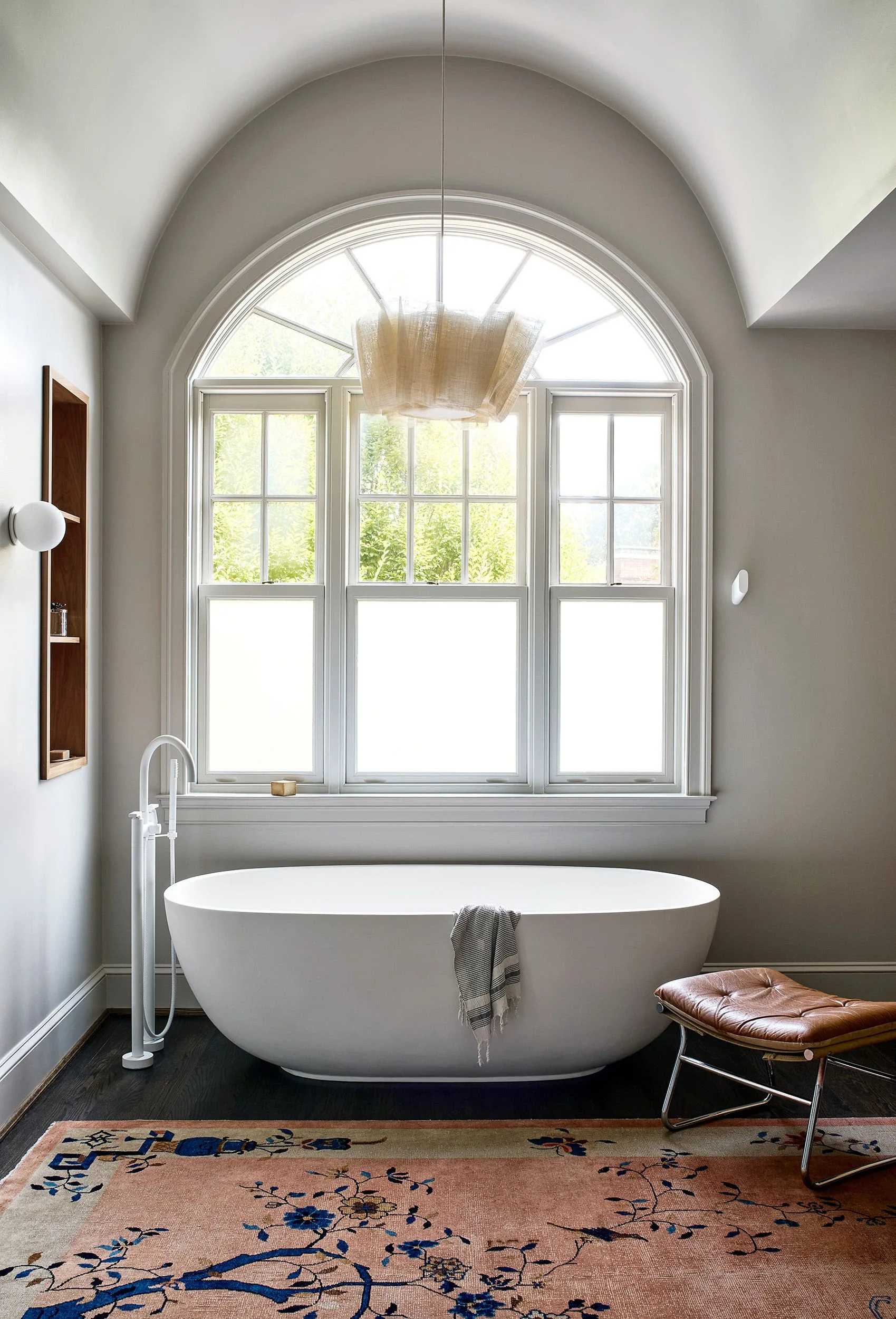 A white freestanding bathtub sits beneath a large arched window in a bathroom featuring a vaulted ceiling, a patterned rug, and a leather stool.