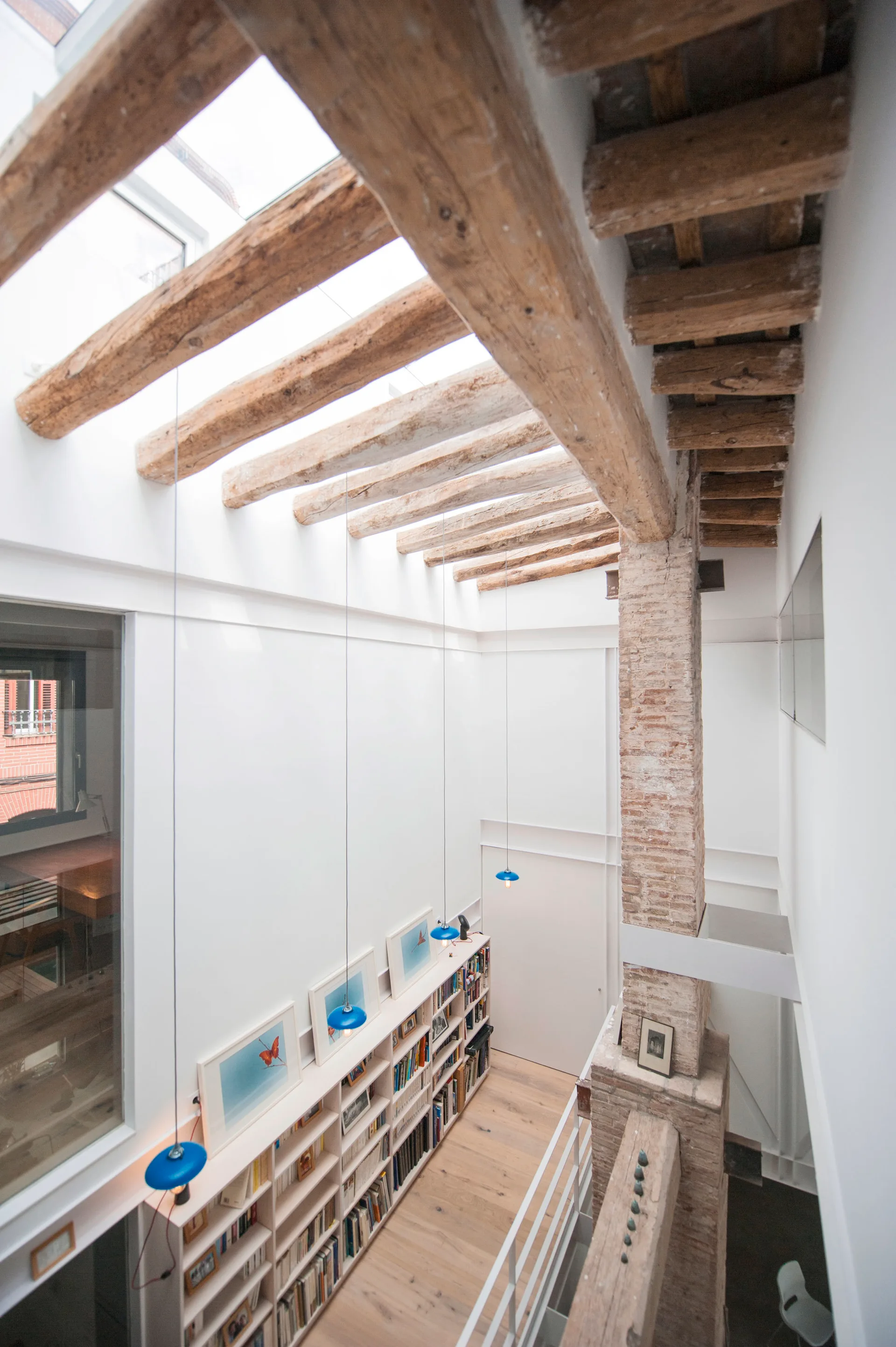 Looking down at a white built-in bookshelf in a loft corridor under a skylight ceiling with exposed wood beams and a brick column.
